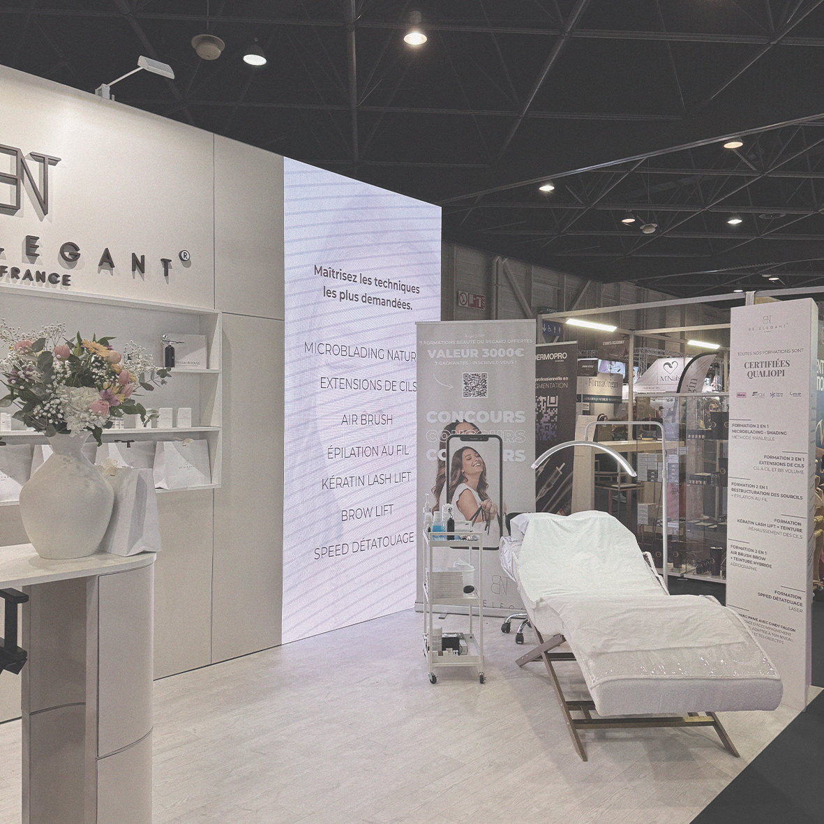 Espace de démonstration de soins de beauté avec un lit de traitement, un présentoir avec des produits cosmétiques, un grand écran affichant la liste de techniques de soins et des affiches promotionnelles.