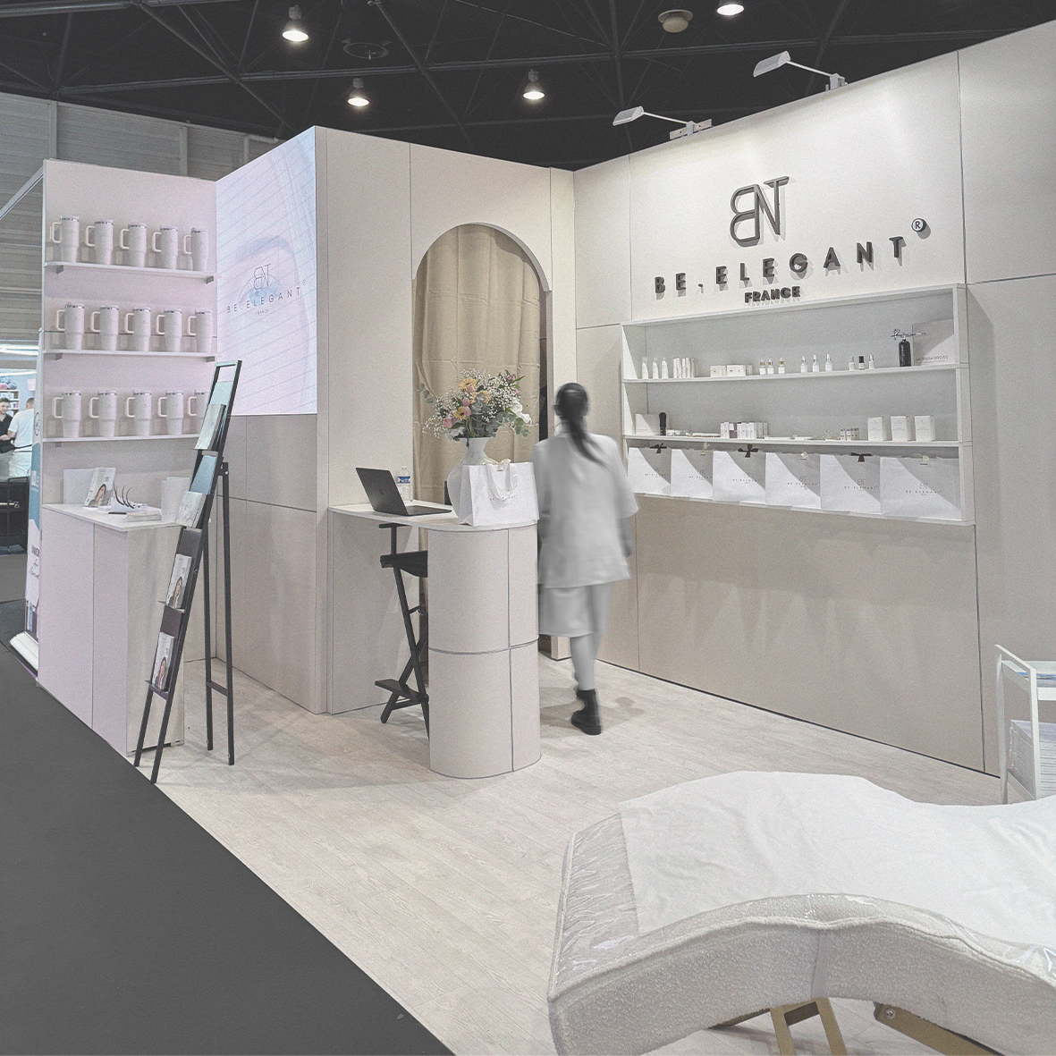 Stand d'exposition de la marque BE. ELEGANT, France, avec étagères de produits blancs, un écran numérique, une table avec un vase de fleurs, et une personne en blanc.