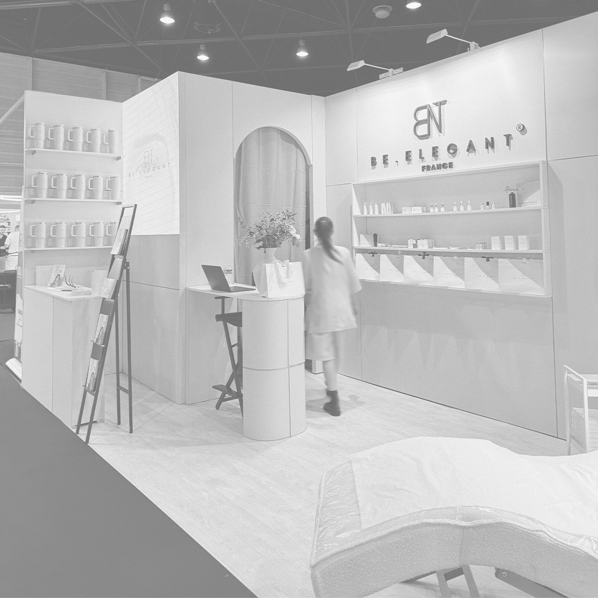 Une personne dans un stand d'exposition blanc avec des étagères contenant des produits, une table avec un ordinateur portable et des fleurs, et un panneau avec le logo 'BE ELEGANT' et 'FRANCE'.