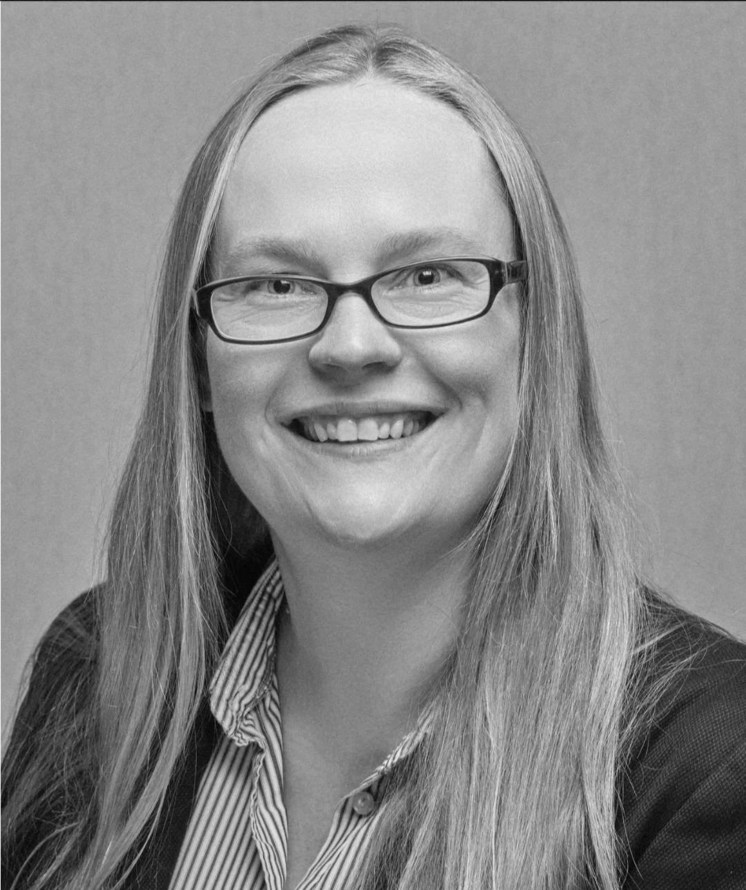 A black and white portrait of a smiling woman wearing glasses, a striped shirt, and a dark blazer.