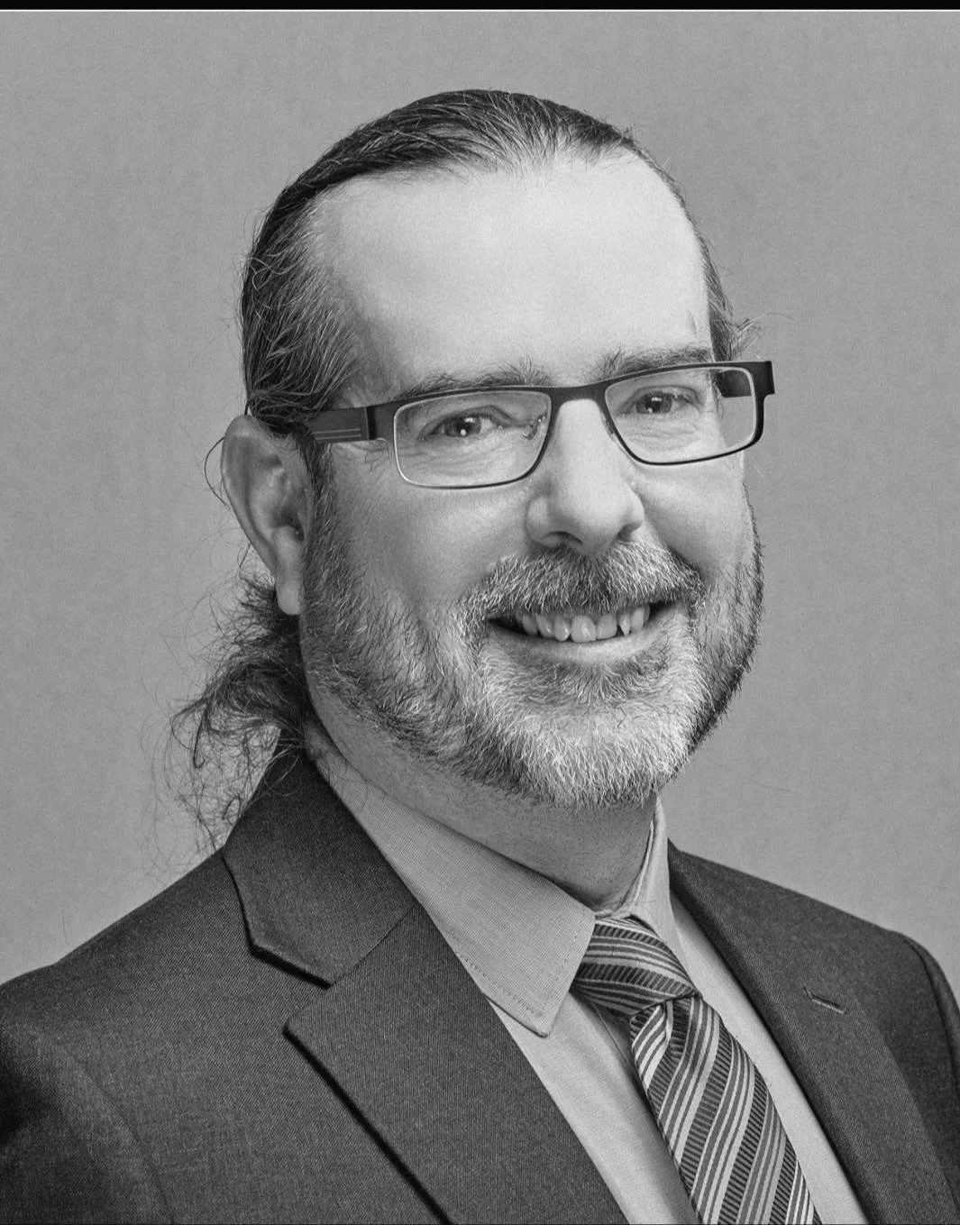 Black and white portrait of a man with glasses, a beard, and long hair tied back, wearing a suit and tie, smiling at the camera.