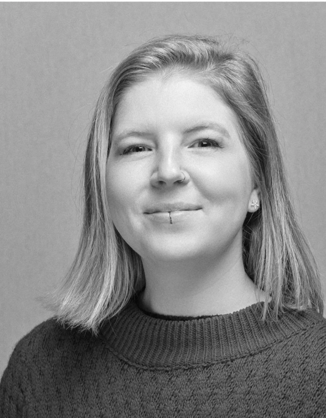 Black and white portrait of a smiling woman with shoulder-length hair, nose piercing, and a lip ring, wearing a dark sweater against a plain background.