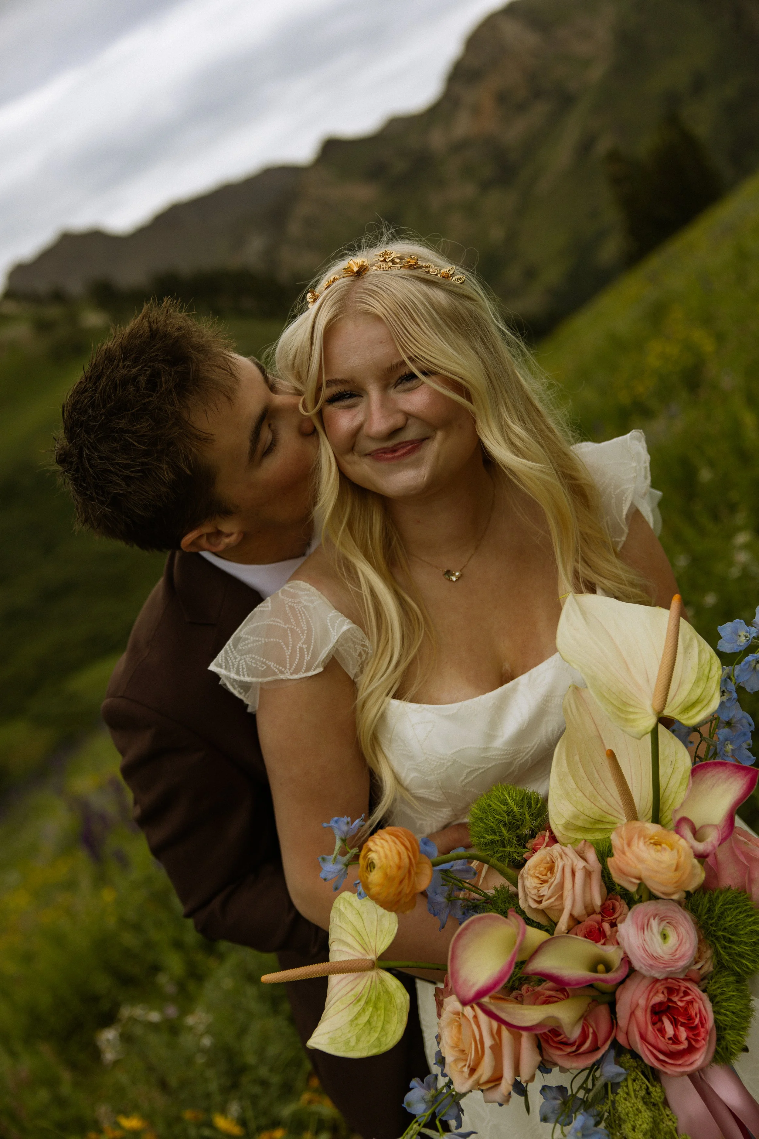 A smiling blonde woman in a white dress holding a large bouquet of colorful flowers with a man kissing her cheek, outdoor mountain landscape in the background.