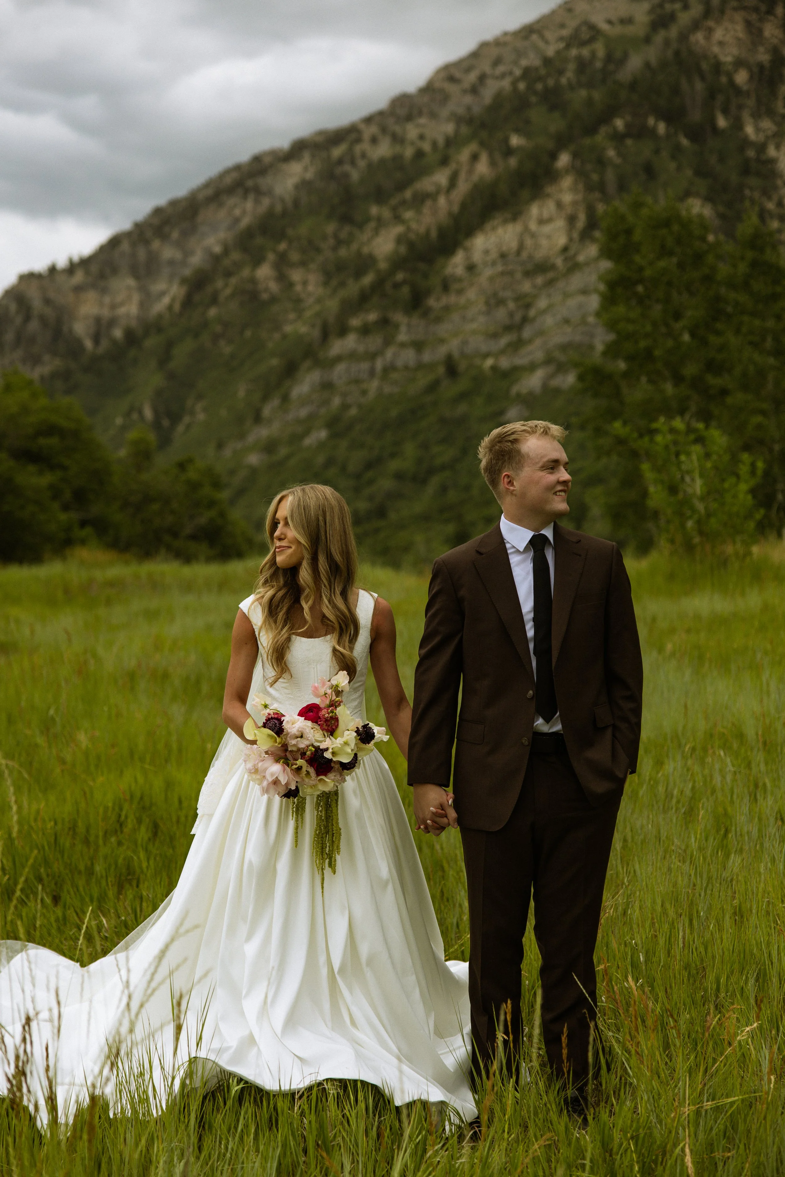 A bride and groom holding hands in a grassy field with mountains in the background, the bride wearing a white wedding dress holding a bouquet of flowers, and the groom in a black suit and tie.
