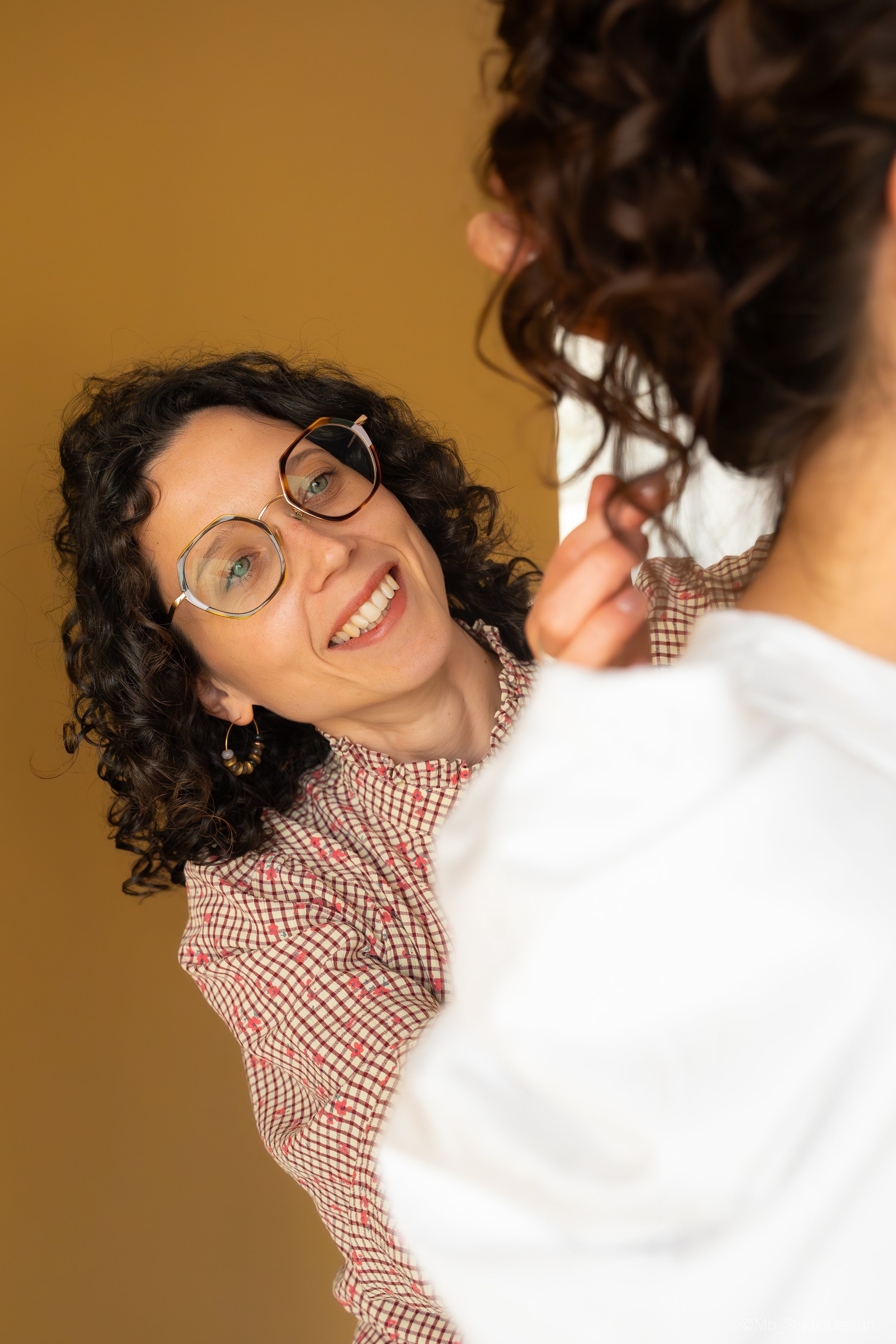 Une femme avec des cheveux bouclés et des lunettes discute avec une autre personne dont on voit seulement la nuque et une partie de l'épaule, dans un décor intérieur aux murs beige.