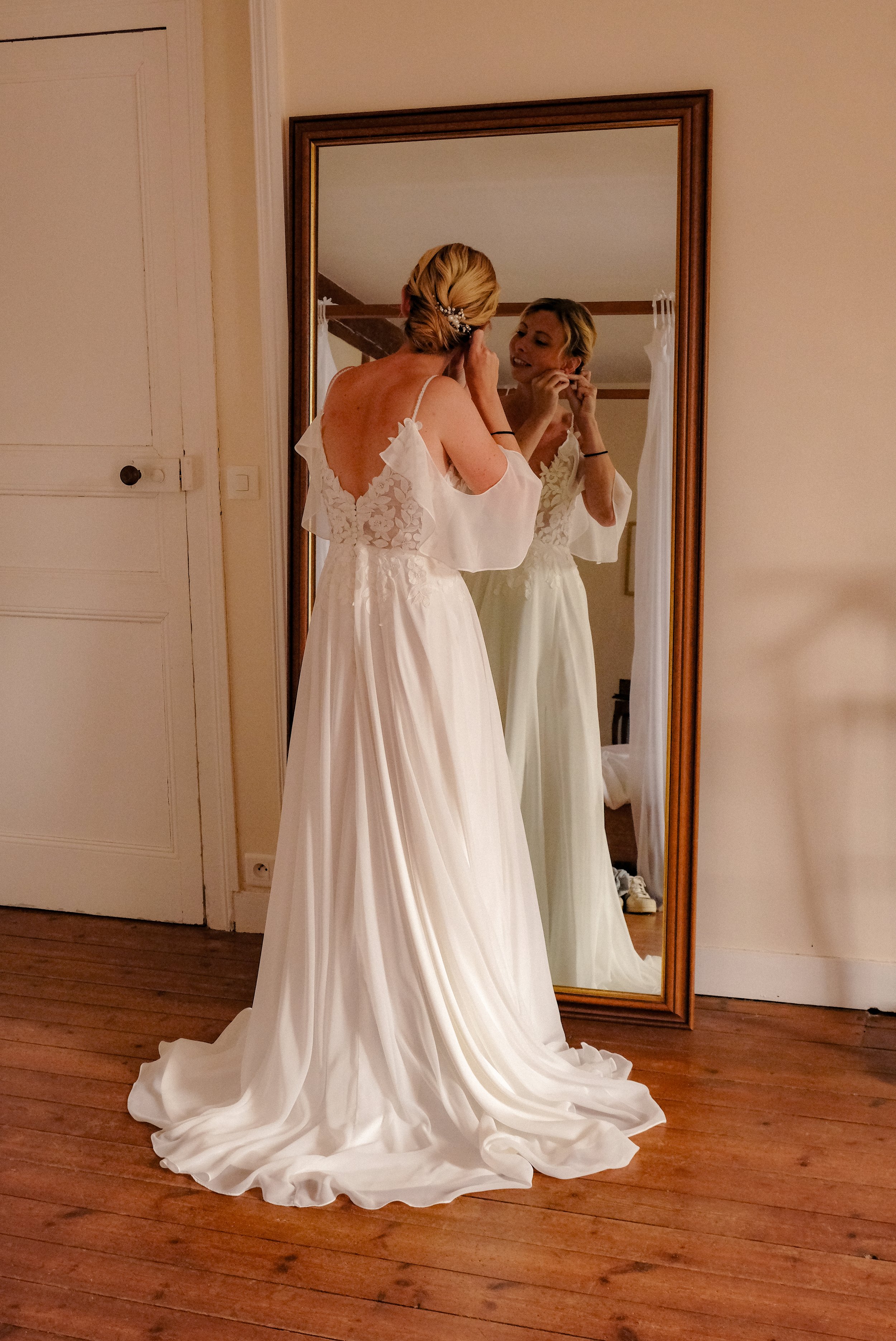 Une femme en robe de mariée blanche devant un miroir, ajustant ses boucles d'oreilles, dans une pièce avec parquet en bois.