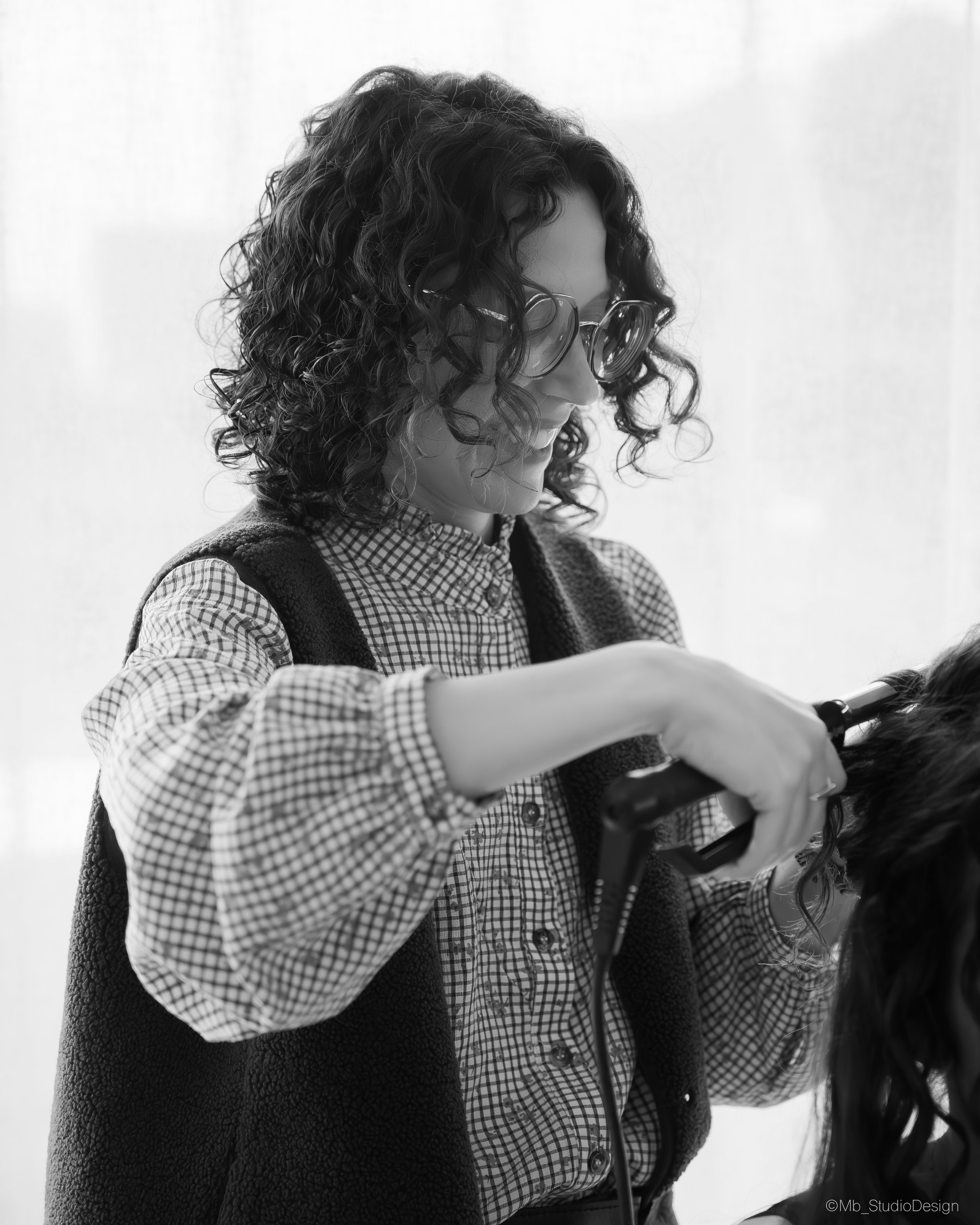 Une femme portant des lunettes de soleil et une veste en laine, tenait une queue de cheval avec un accessoire en métal, en regardant vers le haut. Elle porte une chemise à carreaux et est dans un environnement intérieur avec un fond clair.