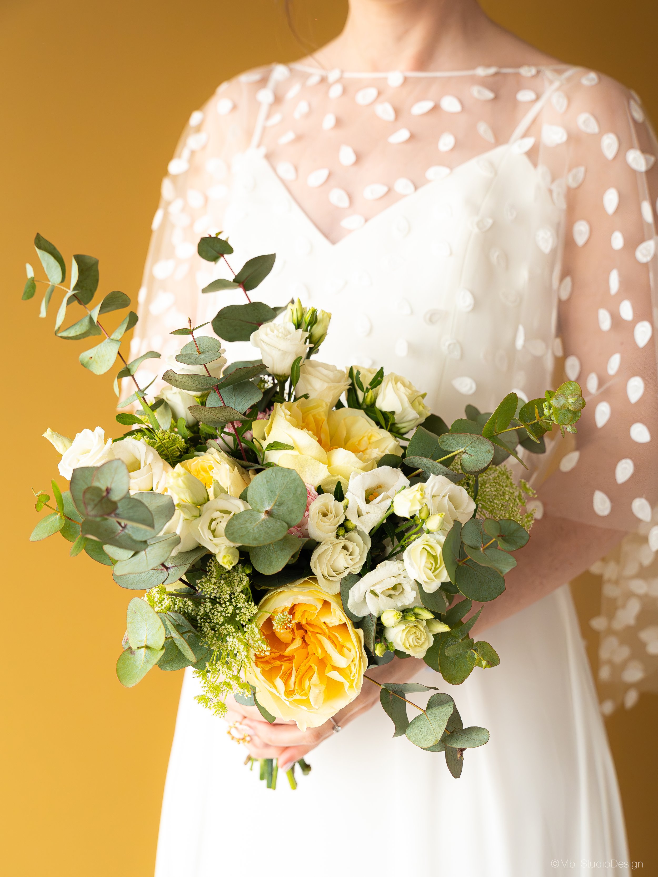 Femme en robe blanche tenant un bouquet de fleurs avec des feuilles vertes contre un fond jaune.