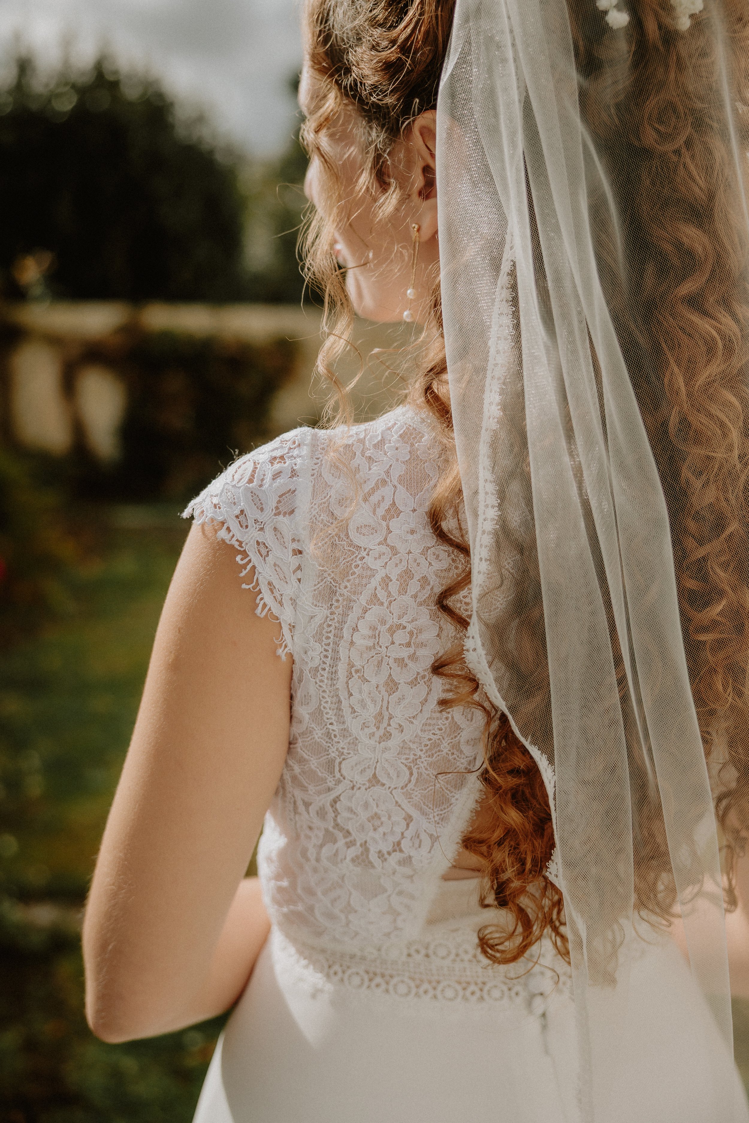 Femme en robe de mariage avec voile en dentelle, vue de côté, dans un jardin en extérieur.