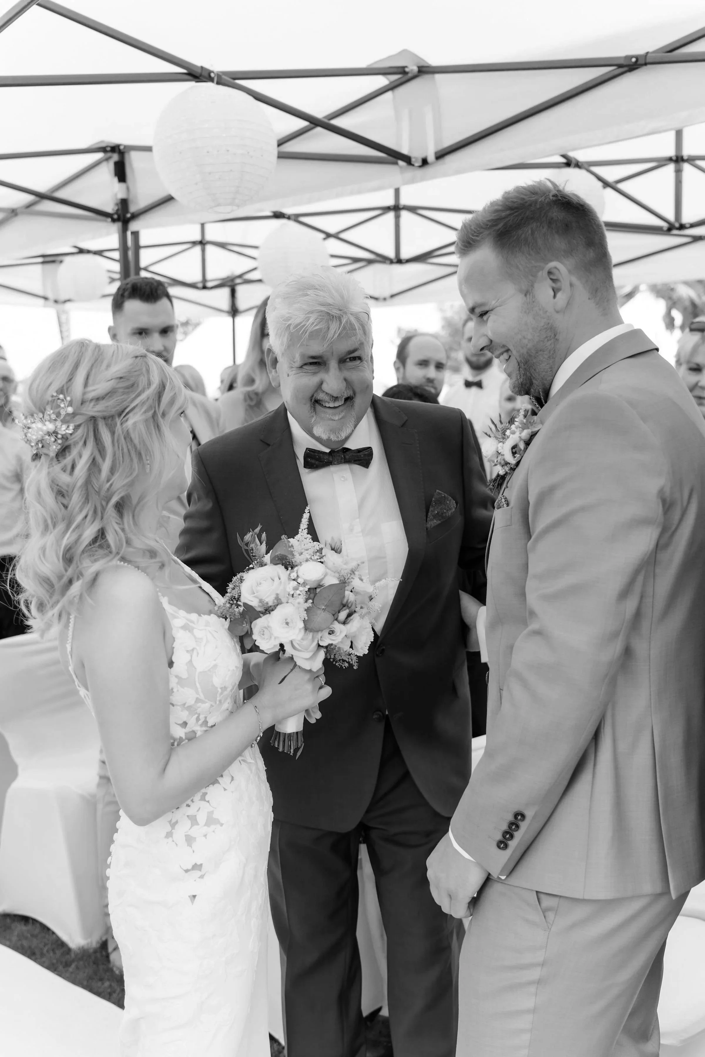 A black and white photo of a wedding ceremony showing a bride and groom holding hands while standing in front of the father of the bride, with guests in the background under canopies decorated with lanterns.