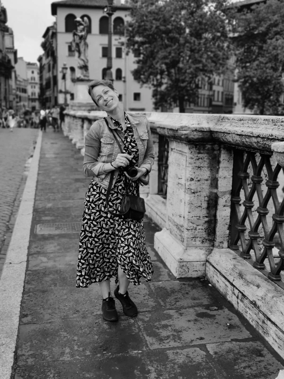 A smiling woman holding a camera and wearing a patterned dress and denim jacket, standing on a bridge with stone and iron railing in a European city.