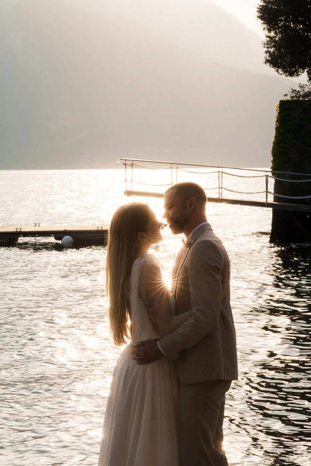 A couple in wedding attire stands close together on a dock by the water during sunset, with mountains and boats in the background.