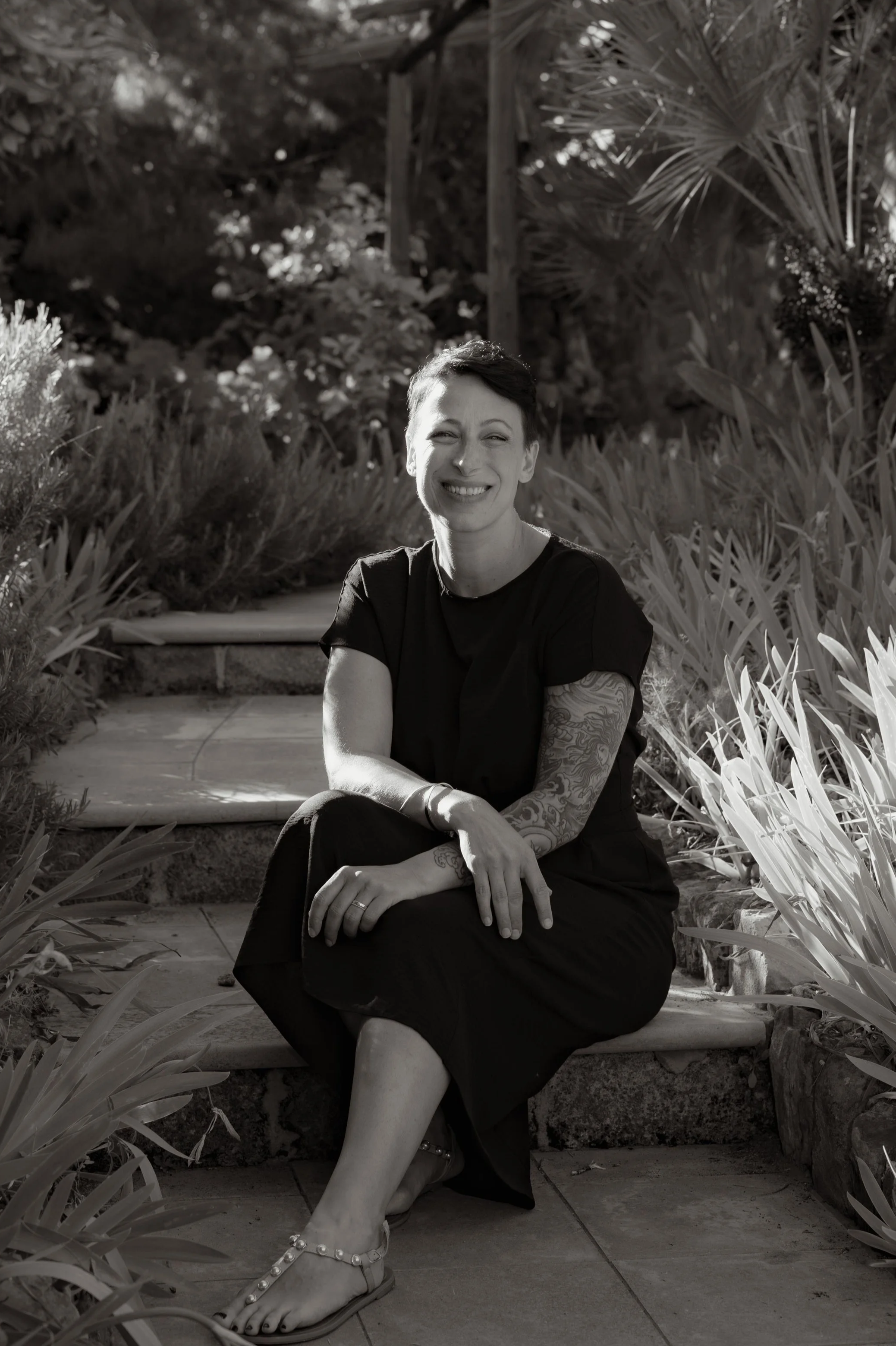Black and white photo of a smiling woman with short hair, sitting on steps outdoors surrounded by plants.