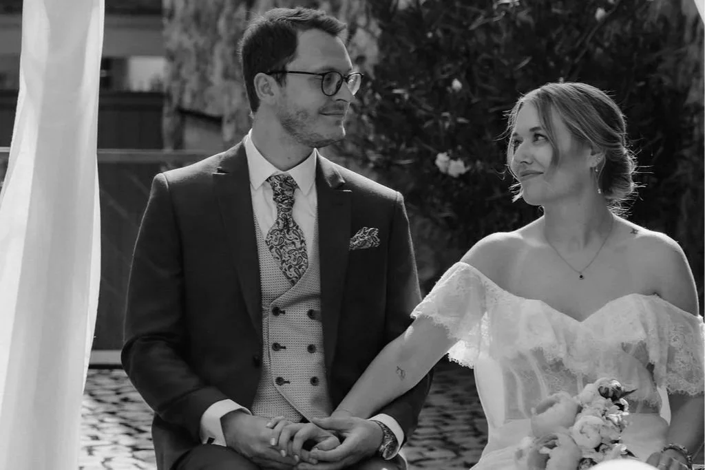 A black and white photo of a couple at a wedding ceremony, holding hands and gazing at each other. The man is dressed in a suit with a patterned tie and glasses, and the woman is wearing an off-the-shoulder lace wedding dress, holding a bouquet of flowers.