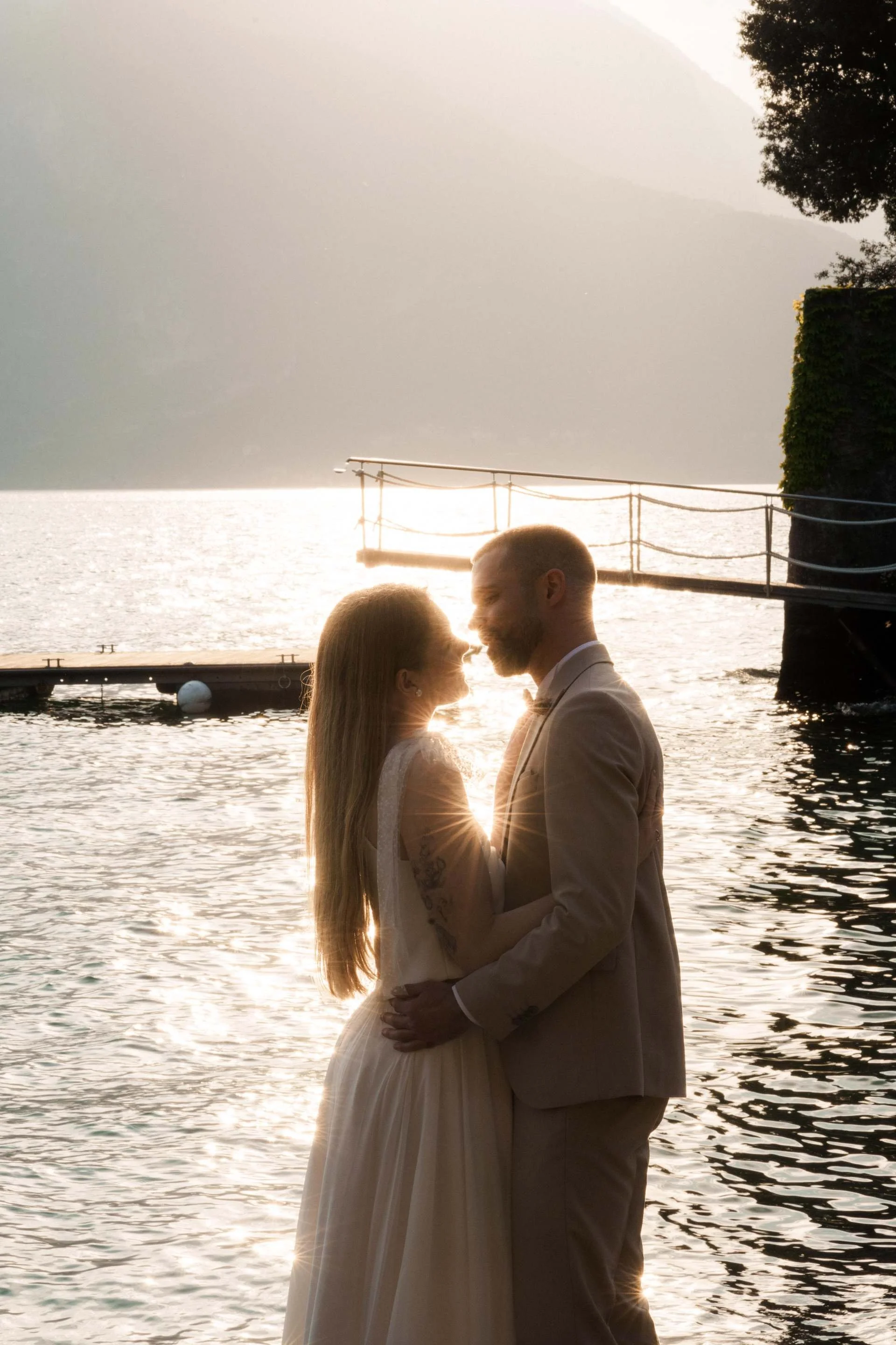 A bride and groom standing close together by a body of water during sunset, with mountains in the background, Varenna, Italy.