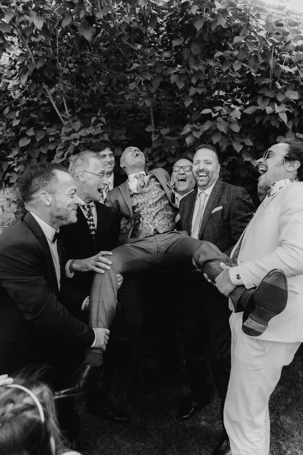 Group of men, dressed in suits and tuxedos, lifting and holding a man dressed in a patterned shirt and plaid pants, all laughing joyfully in front of a leafy backdrop.