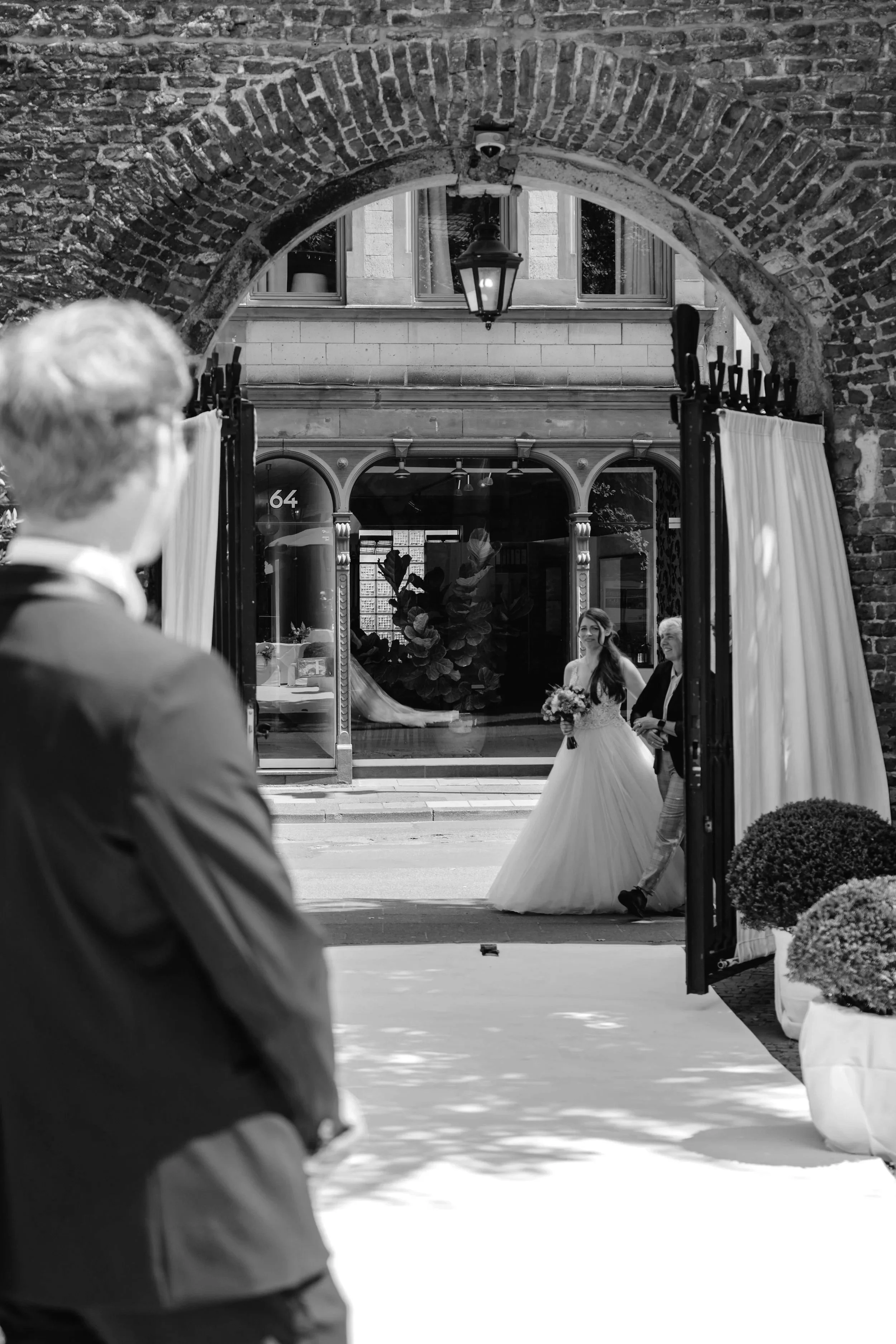 A bride holding a bouquet walks into a building through open curtains, while a person stands outside observing. The scene is viewed through a large archway in a black-and-white photo.