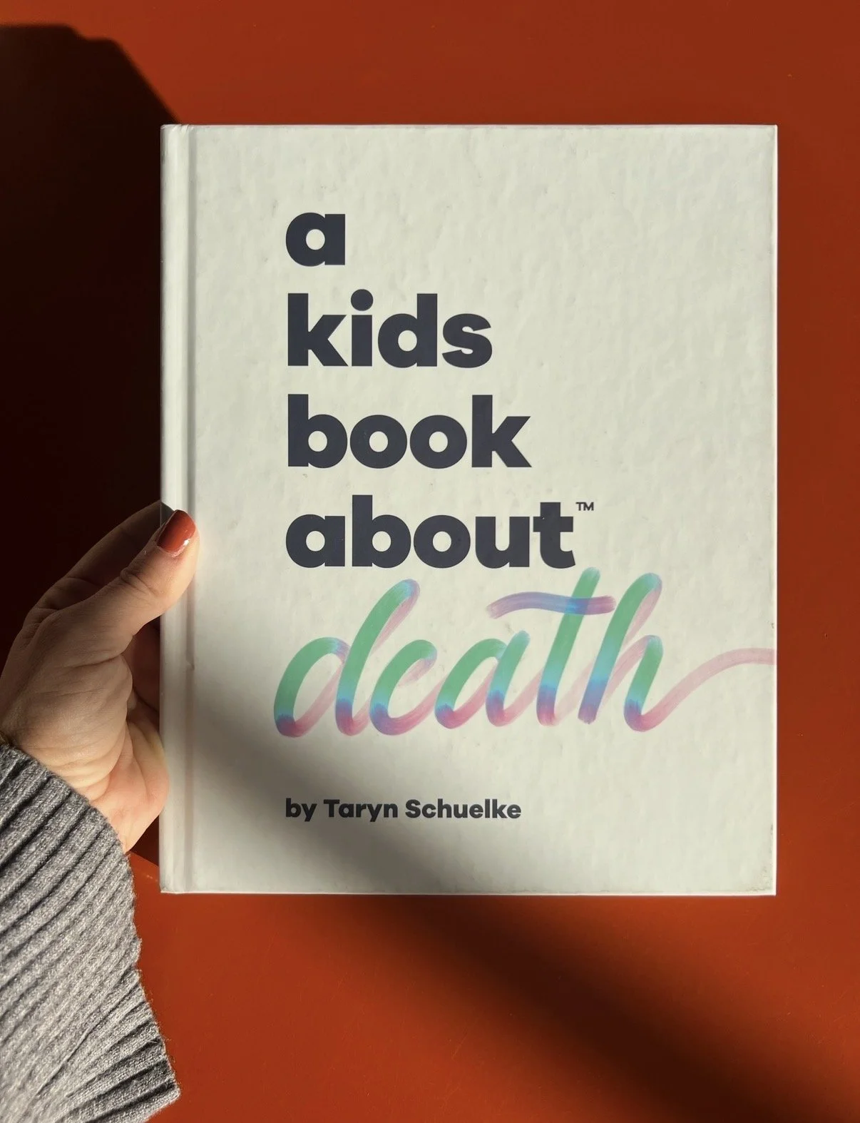 Libro infantil titulado "A Kids Book About Death" de Taryn Schuelke, con letras grandes y coloridas en la portada, sostenido por una mano con esmalte de uñas naranja, contra fondo naranja.