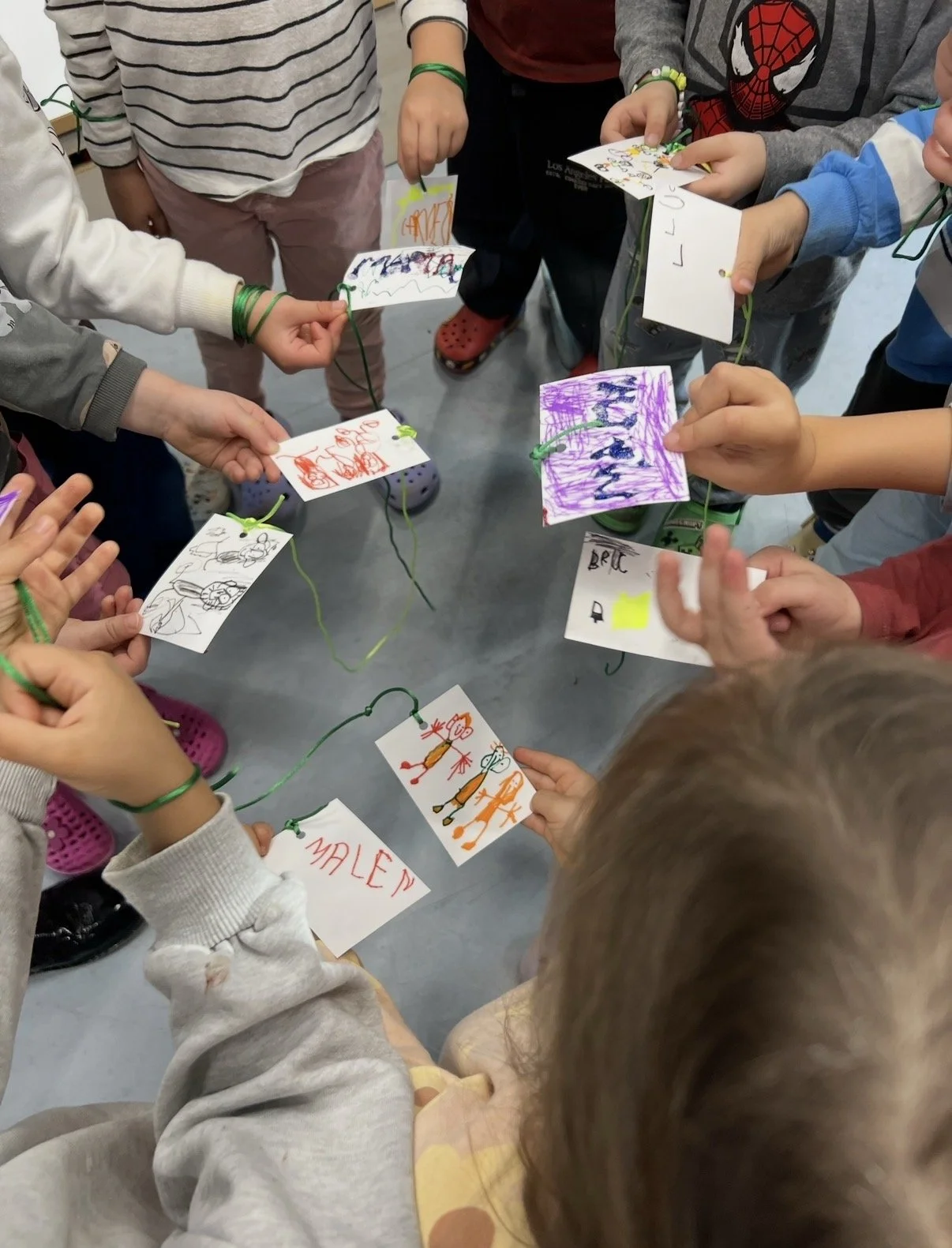 Grupo de niños pequeños muestran dibujos coloridos y notas de papel conectadas con cuerda verde, en un entorno de interior.