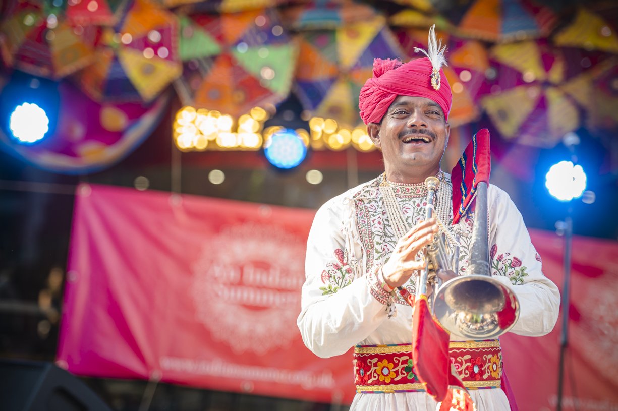 A man in traditional Indian attire, including a pink turban with a feather, is smiling and playing a trumpet, with colorful umbrellas decorating the background at a festive event.