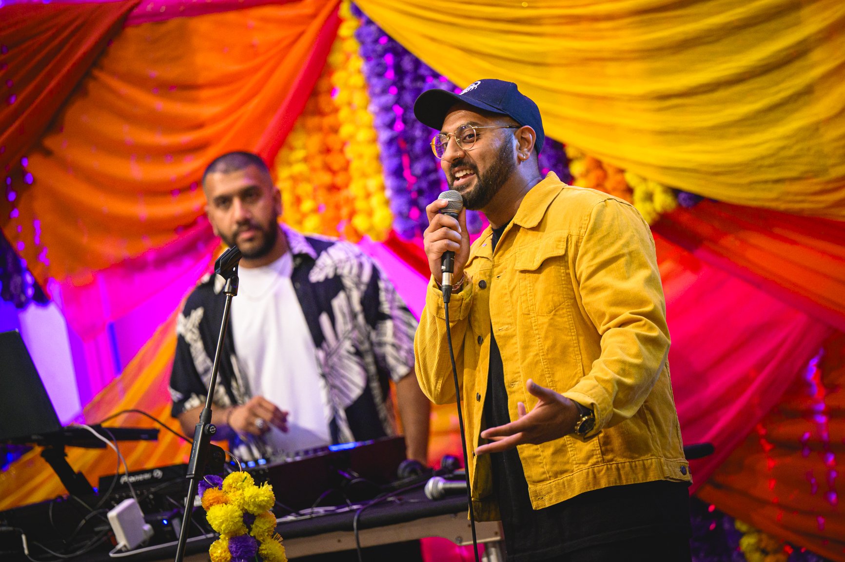 Two men performing on stage with vibrant orange, pink, and purple floral backdrop; one in a yellow jacket and cap singing into a microphone, the other in a patterned shirt with a keyboard and sound equipment nearby.