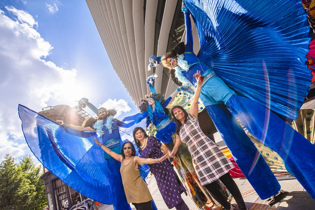 People standing under a large, colorful parade float with blue costumes and whimsical decorations, celebrating outdoors in bright sunlight.