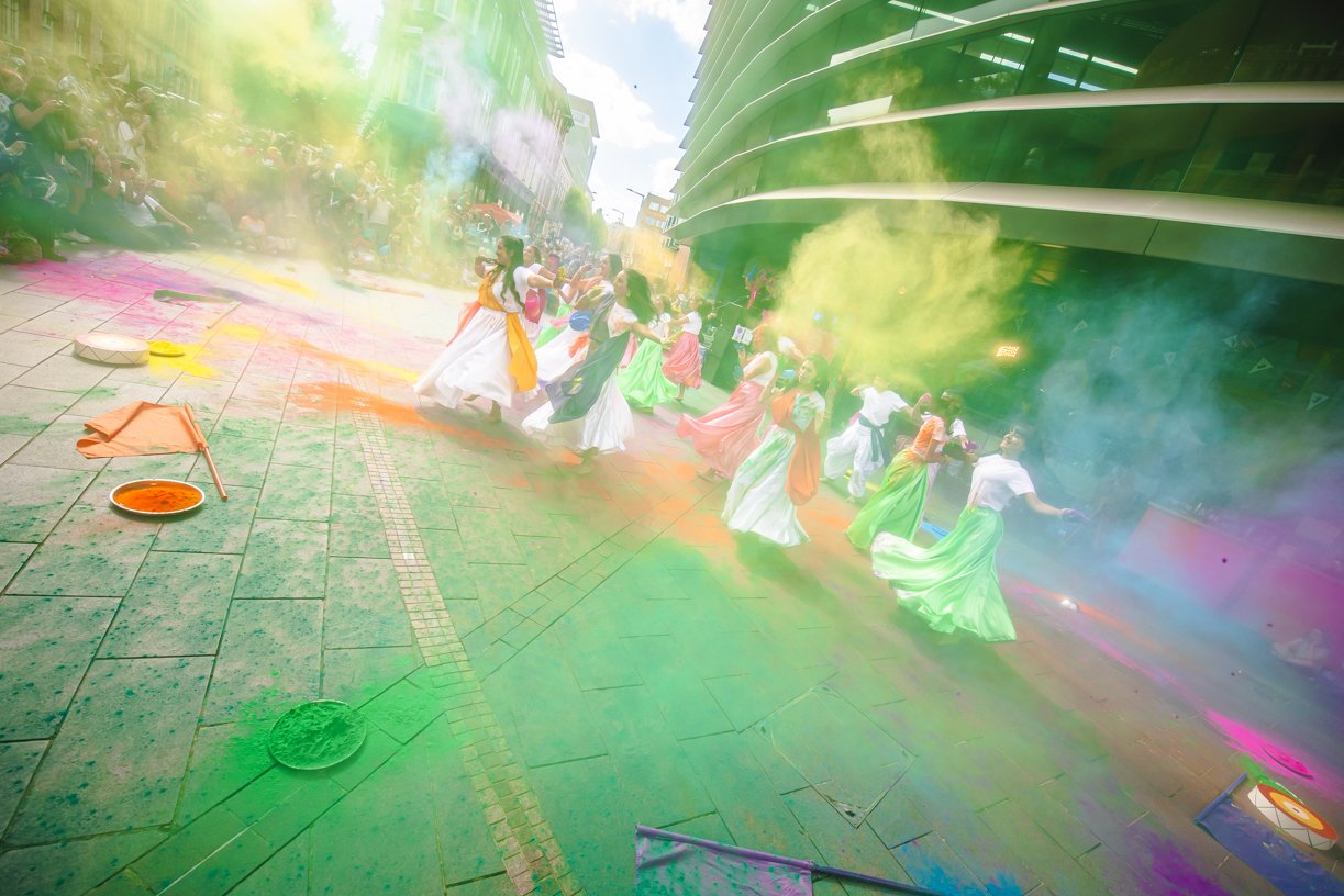 People celebrating with colored powders during a festival on a city street, with buildings in the background and colorful haze in the air.