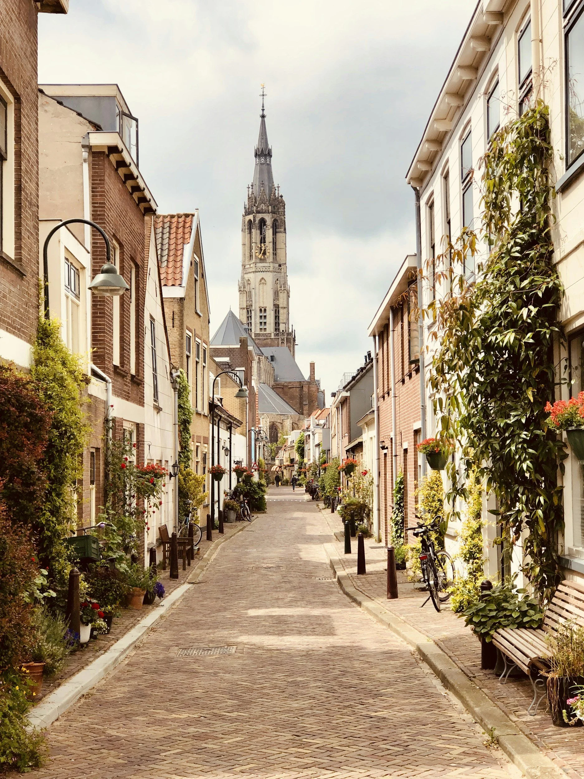 A quaint European street lined with brick and white residential buildings, decorated with potted plants and hanging flower baskets, with a church steeple visible in the background under a cloudy sky.