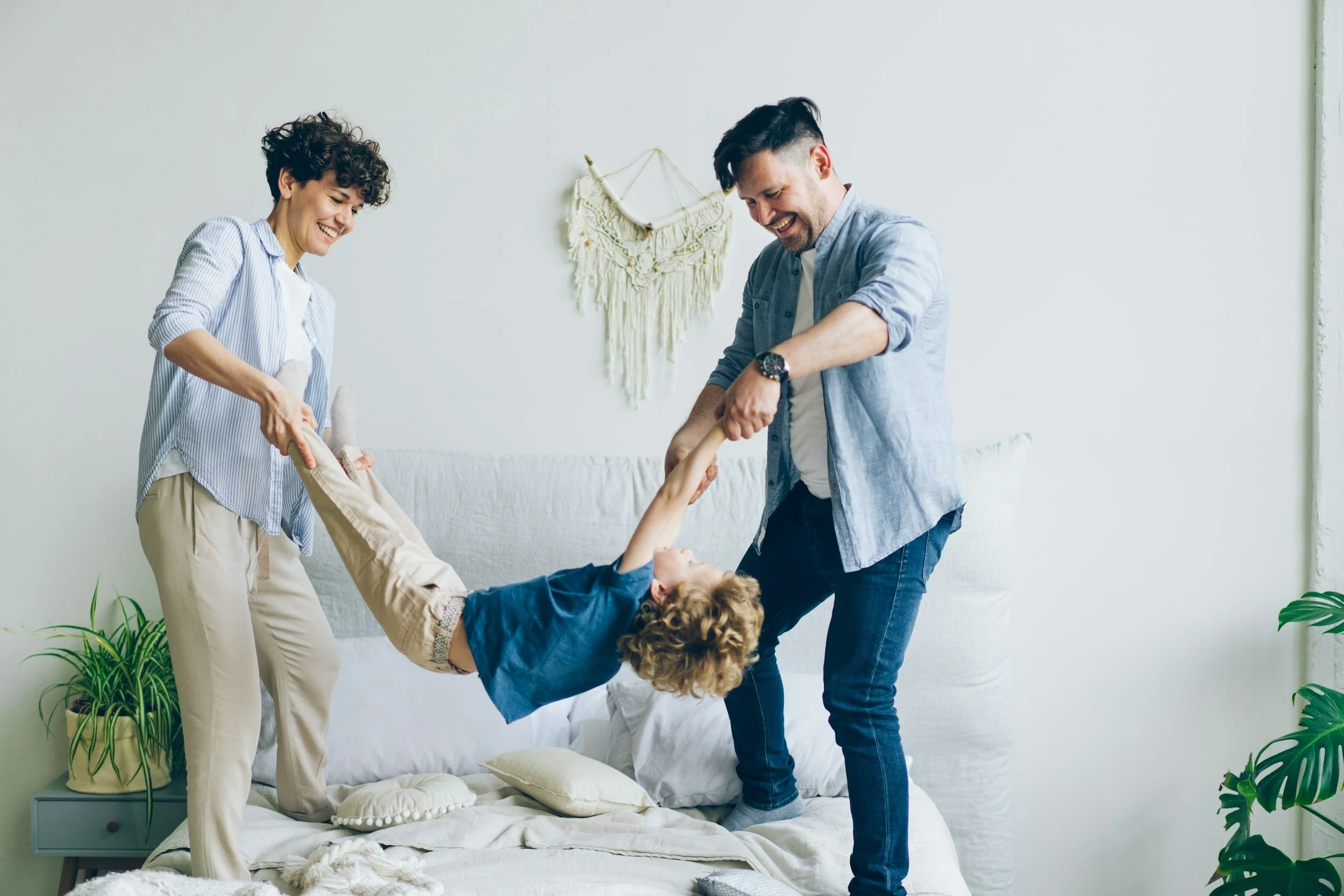 Two adults and a child playing together in a bright living room, with the child hanging upside down from the adults' hands, smiling and enjoying the moment.