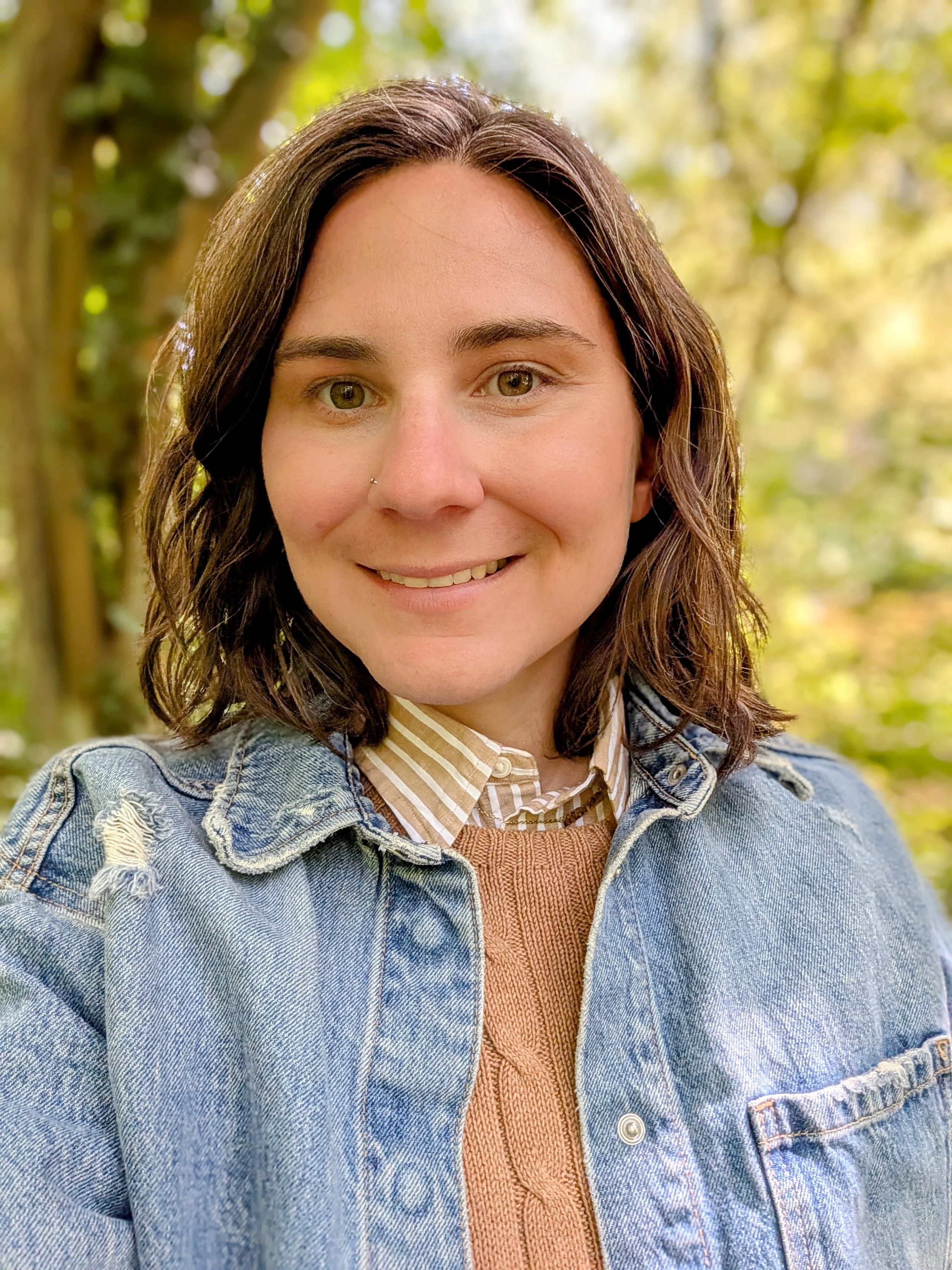 Close-up of a smiling woman with brown hair, wearing a denim jacket over a striped shirt and a rust-colored sweater, outdoors with a background of green trees and sunlight.