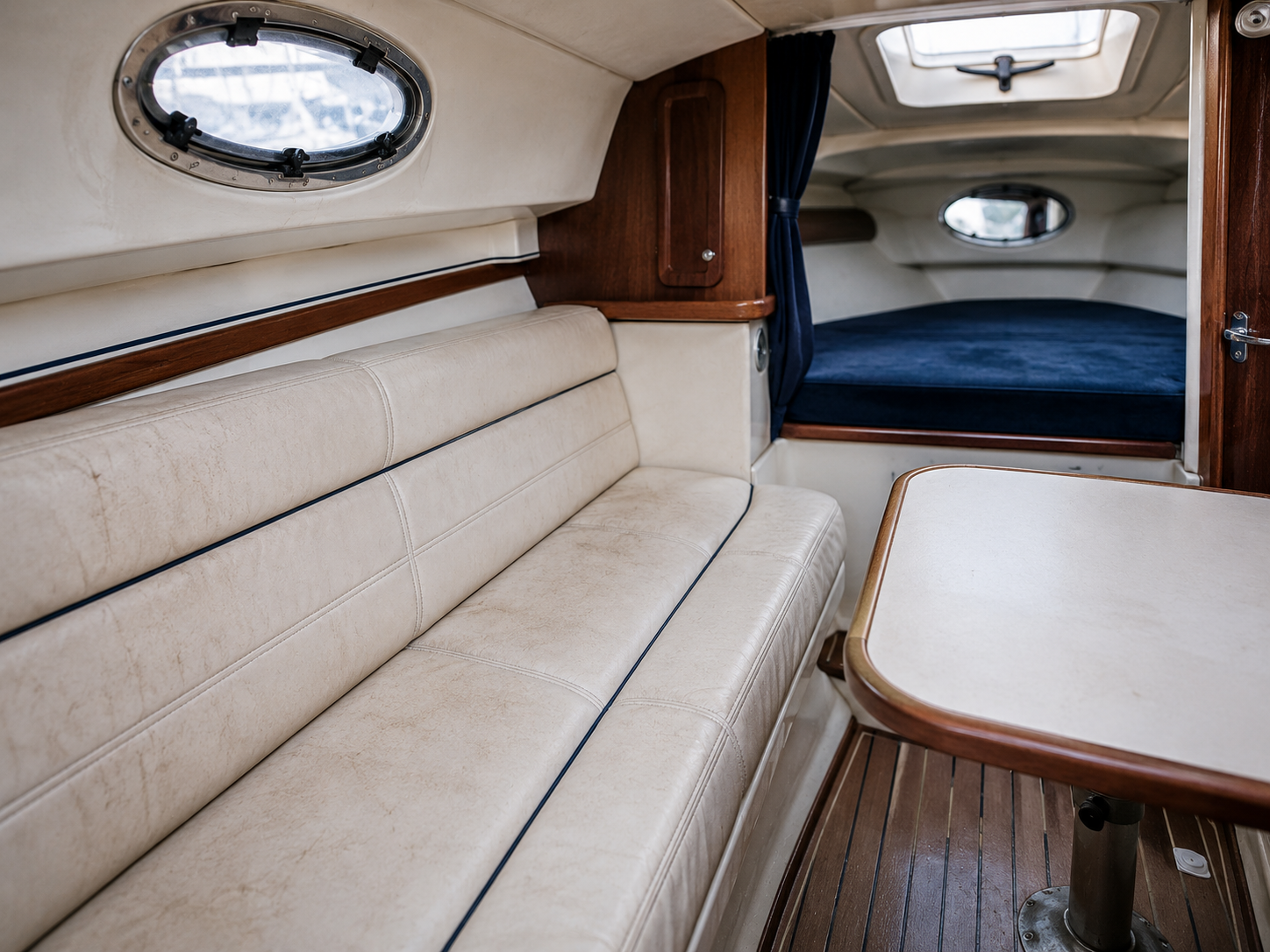 Intérieur d'un bateau avec un canapé en cuir beige, une table en bois, une cabine couchette avec un lit en tissu bleu foncé, fenêtres ovales et un plafond avec hublots.