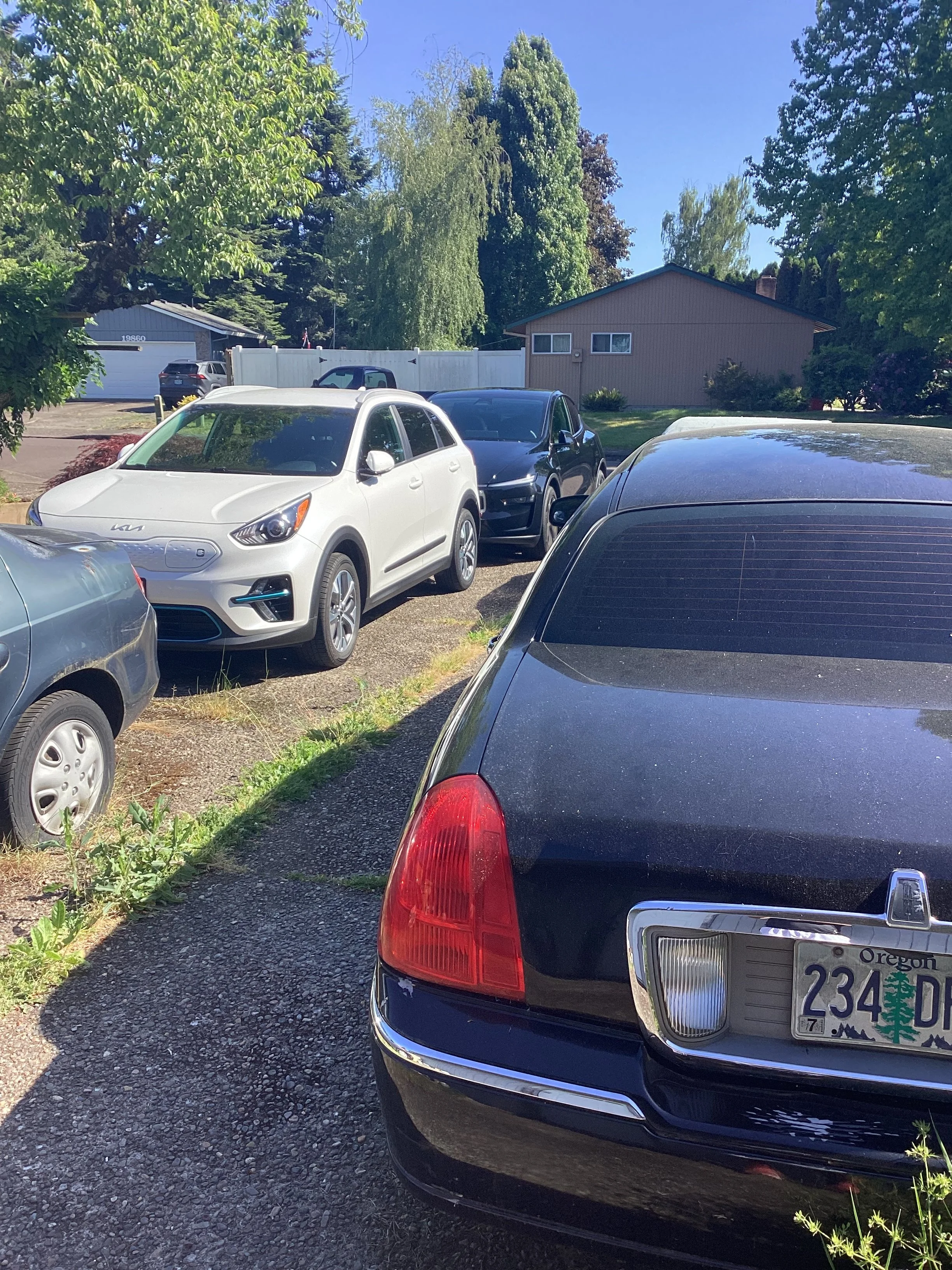 A black Lincoln town car , Tels Y model and Kia electric parked in a driveway with an Oregon license plate, with other cars and a house in the background amidst trees and greenery on a clear sunny day.