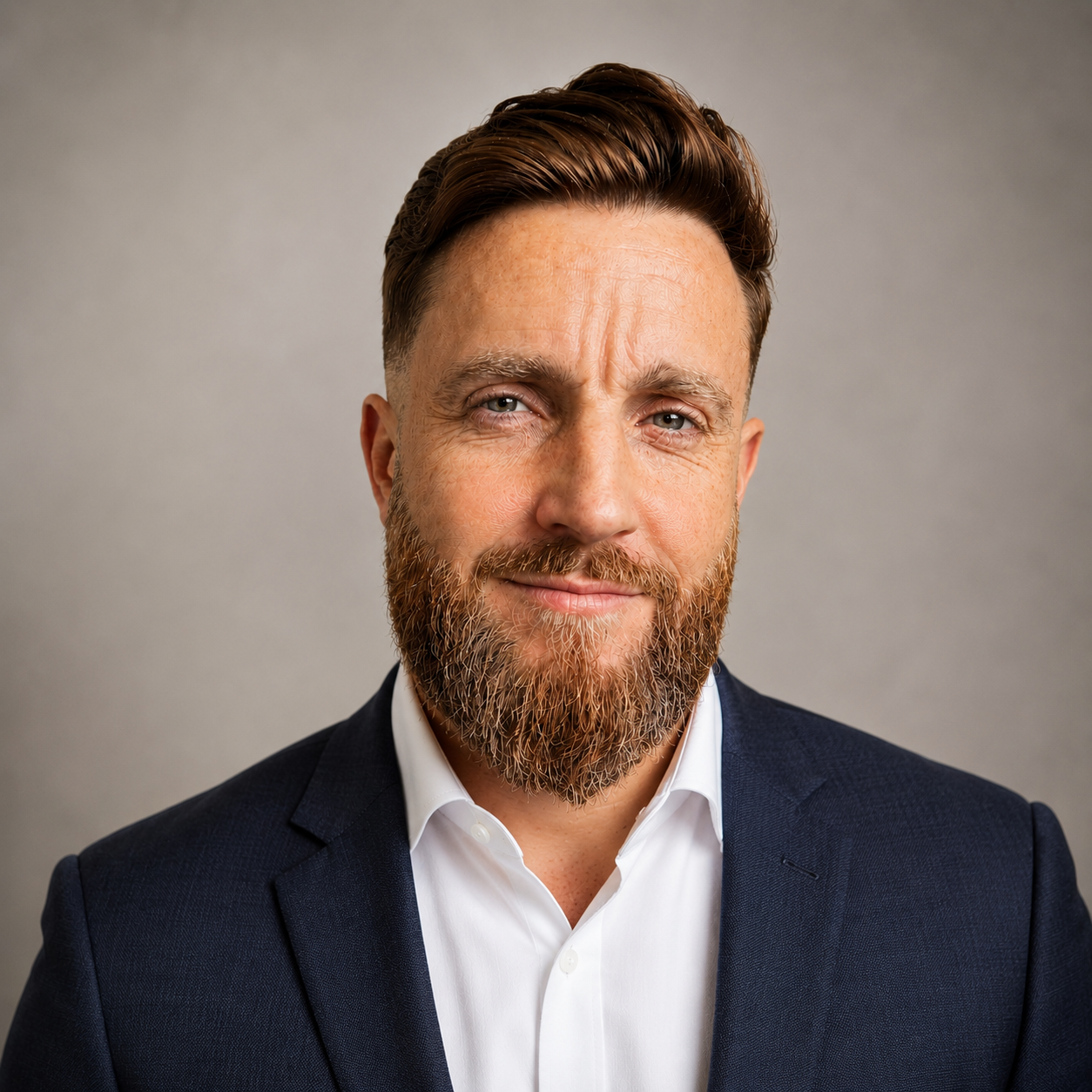 Portrait of a man with a beard, wearing a dark suit and white shirt, smiling against a plain background.