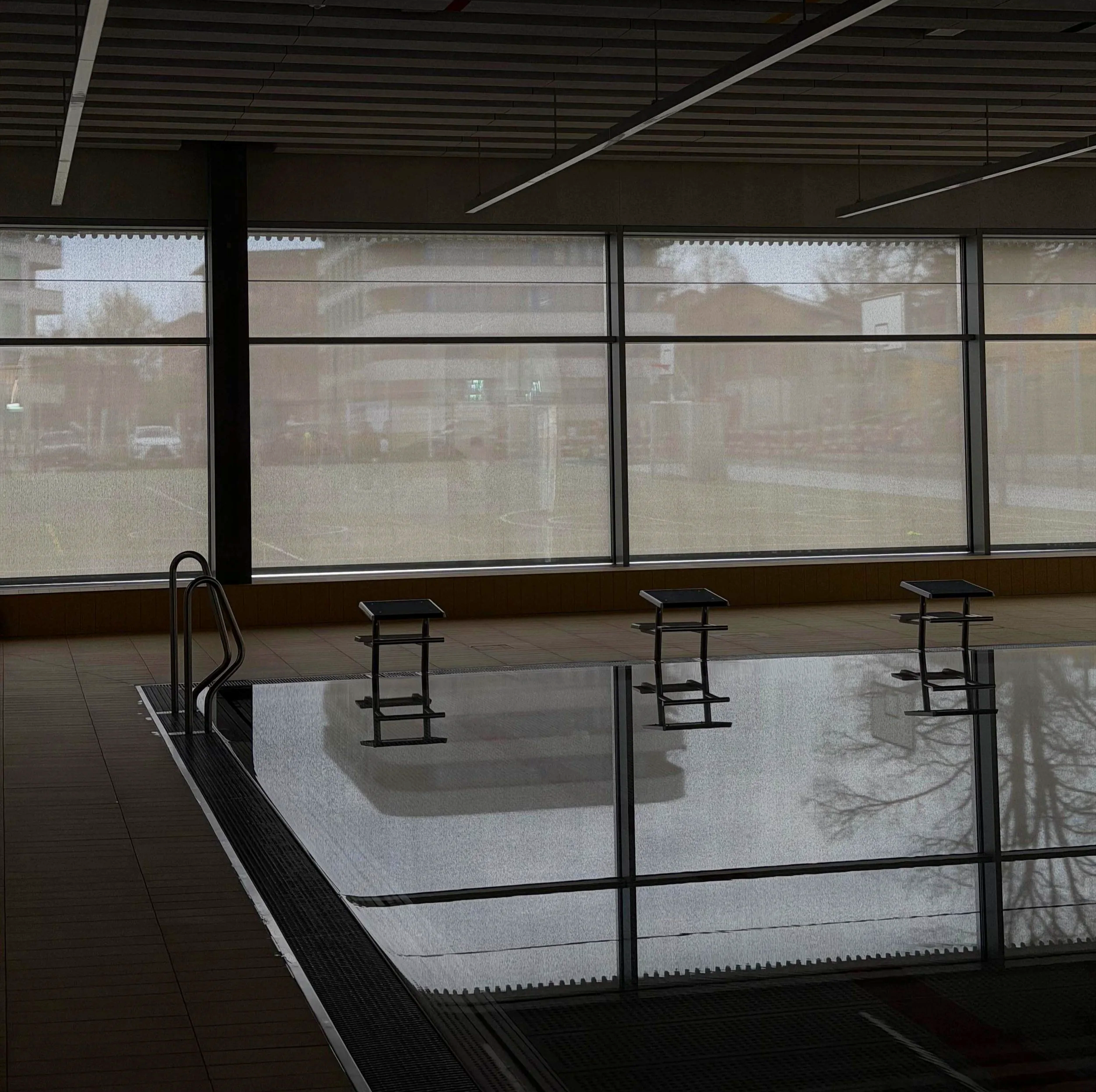 Interior view of a building with large windows, a pool ladder, and mock lounge chairs, possibly a gym or swimming pool area.