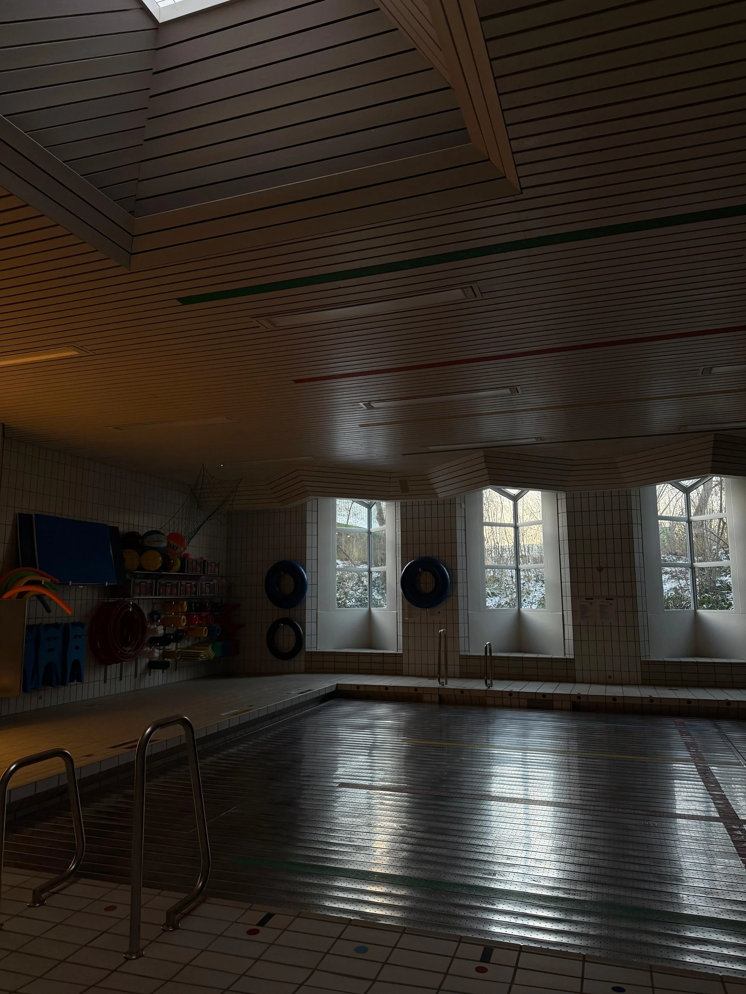 An indoor swimming pool area with large windows, wooden ceiling, and swimming equipment such as kickboards, hula hoops, and balls stored on shelves. The pool has a metal ladder and the floor has white tiles with colored markers.