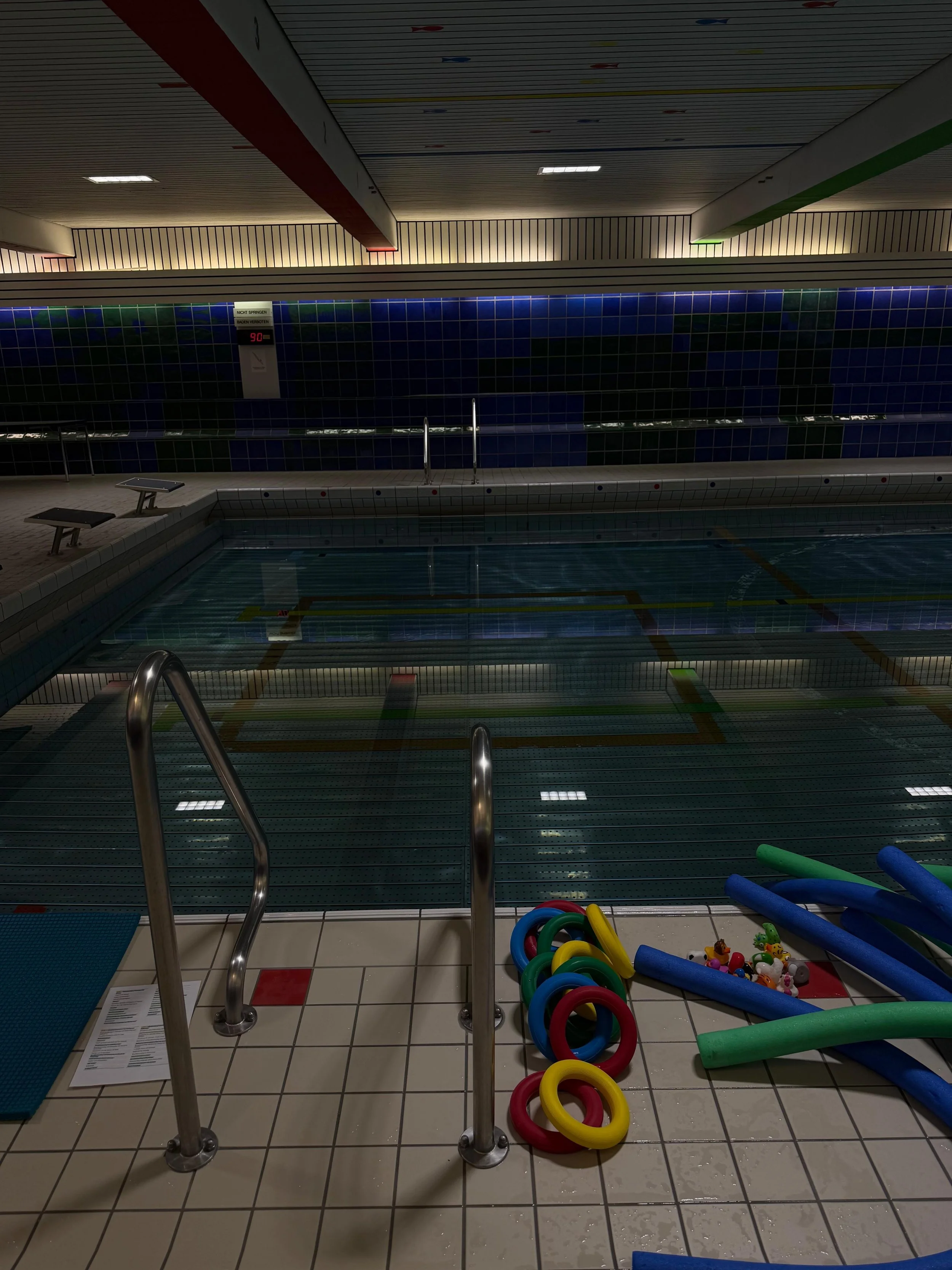 Indoor swimming pool area with a shallow children's pool, colorful pool toys like foam noodles and rings, and water safety equipment; tiled floor and a monitored pool temperature display.