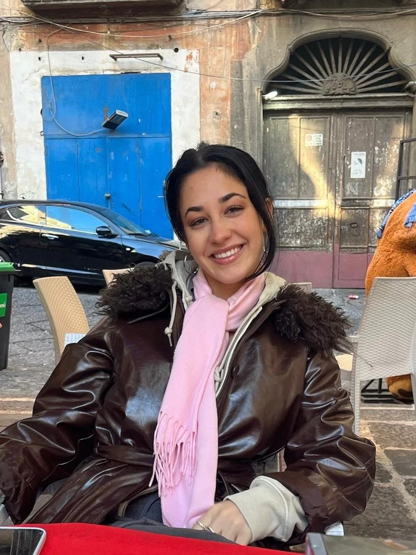 A young woman with dark hair smiling, wearing a brown leather jacket with a fur-lined hood and a pink scarf, seated outdoors at a table.