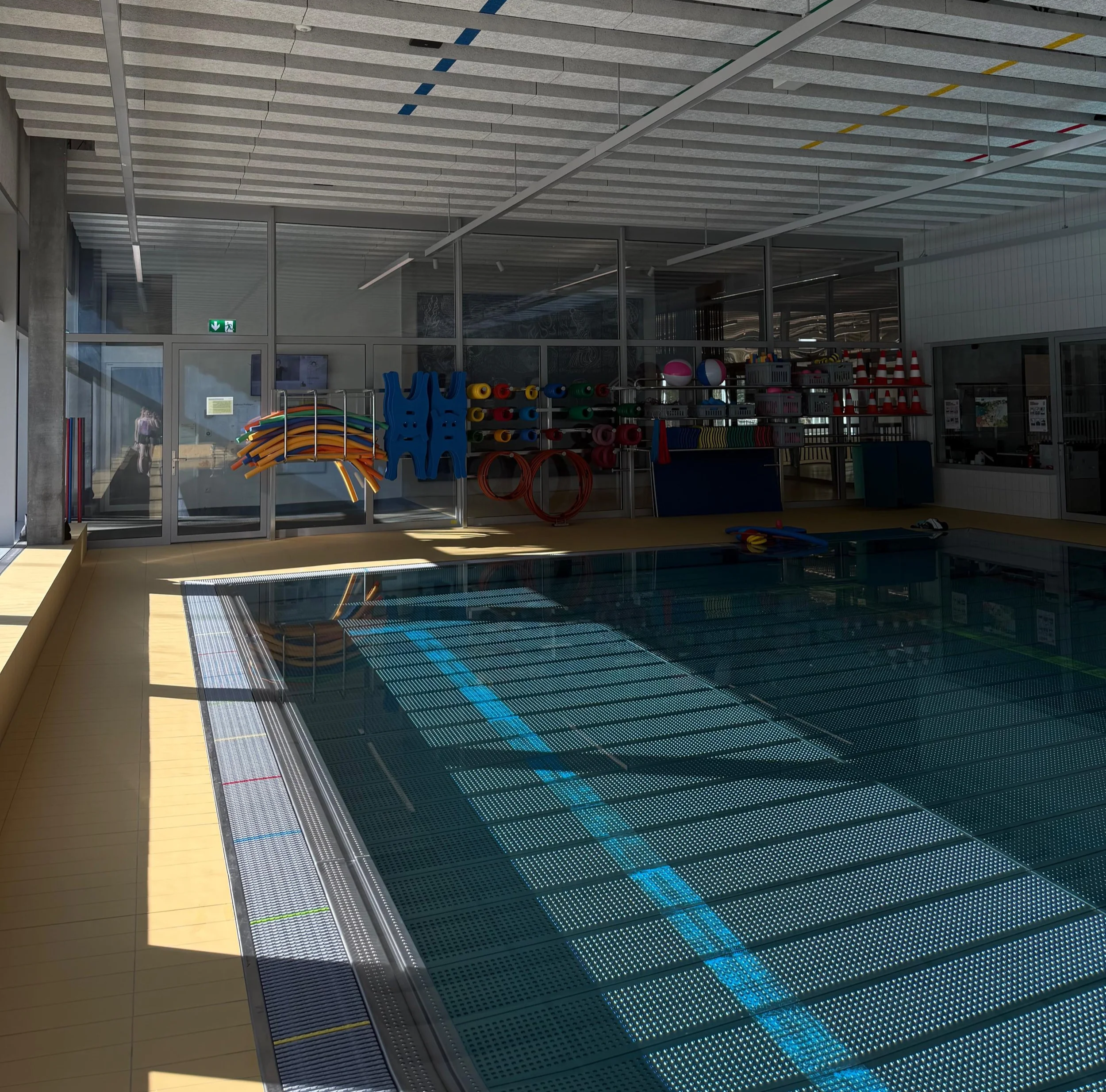 Indoor swimming pool with colorful foam pool noodles and pool balls stored on shelves, glass walls, and water reflecting sunlight.