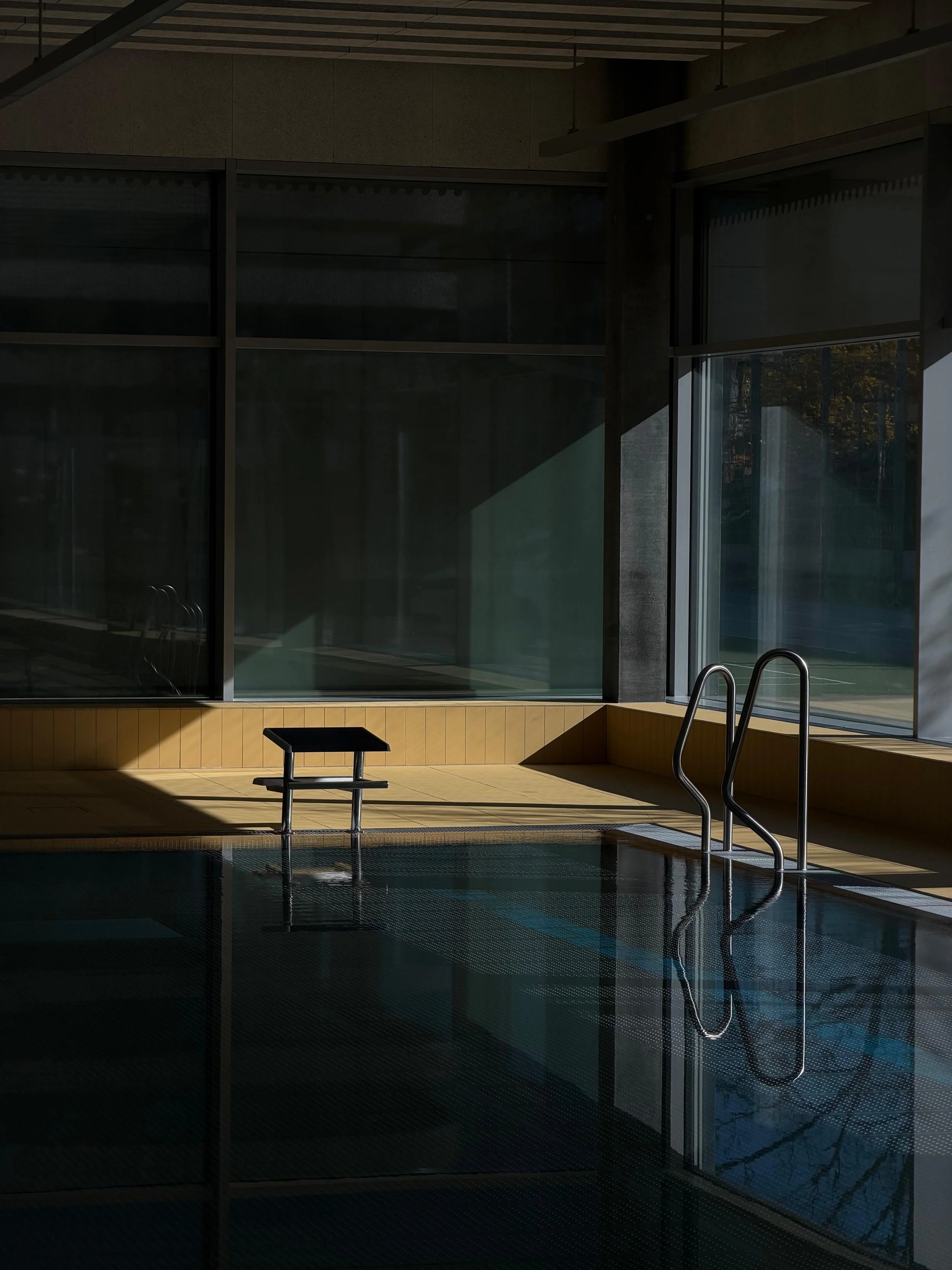 An indoor swimming pool with large windows, a start block, pool ladder and sunlight casting shadows across the tiled deck and water.