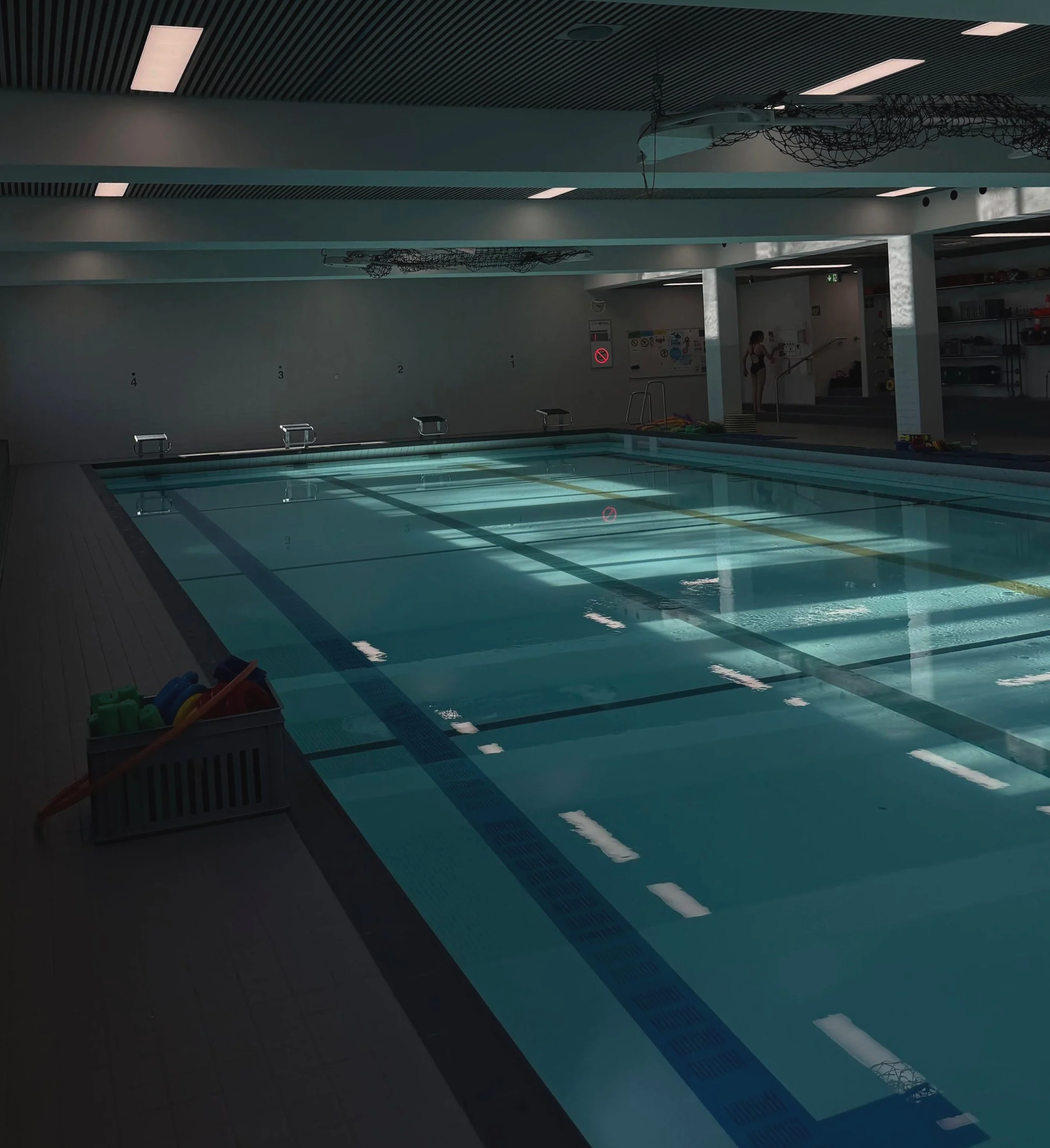 Indoor swimming pool with lane dividers, starting blocks, and a woman in a swimsuit near the stairs. Pool features a modern ceiling with light fixtures, and storage shelves on the side.