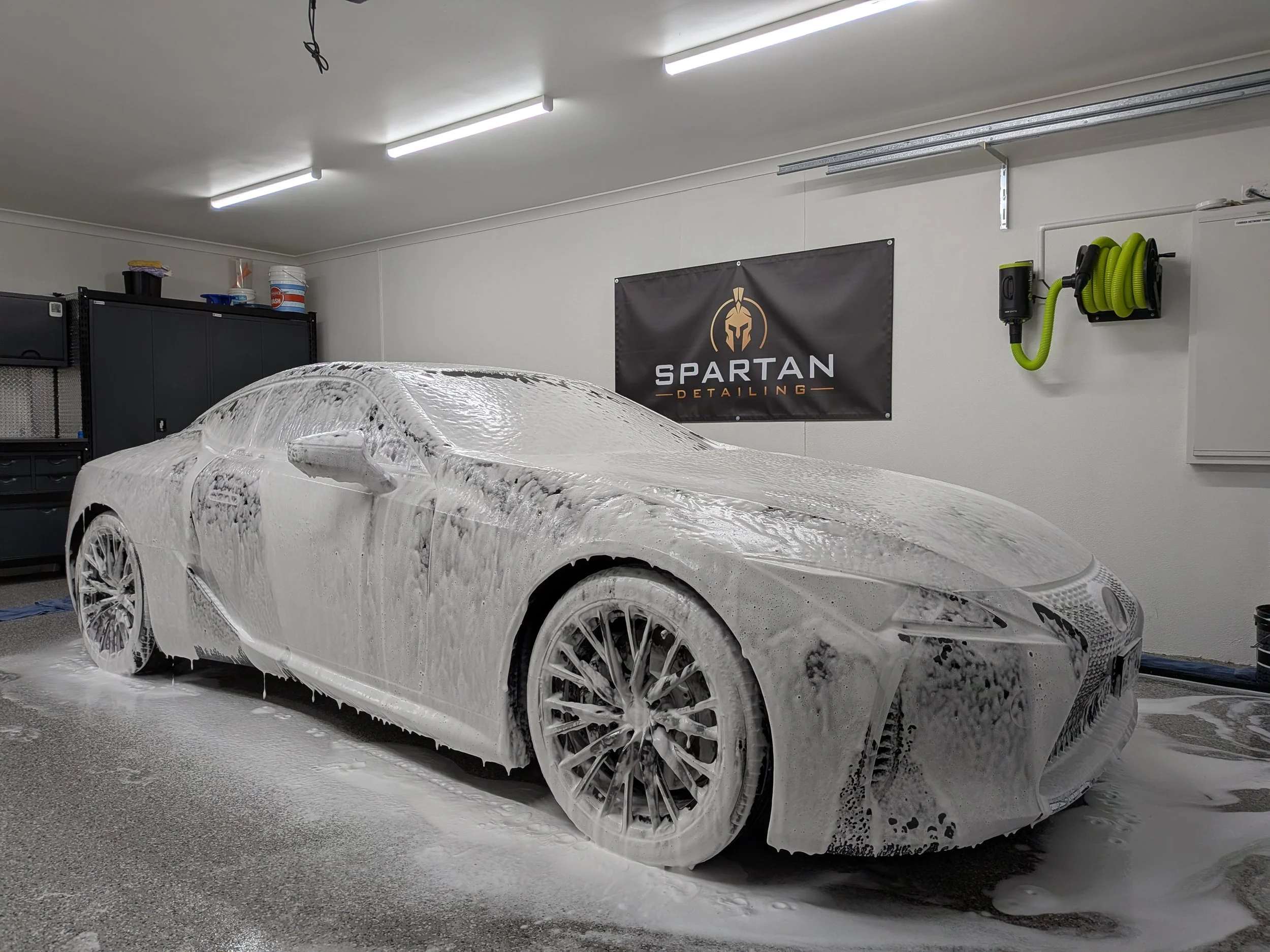 A black Lexus LC500 GT covered in soap suds and foam during a car wash, inside a garage workshop with overhead LED lighting with detailing equipment on the wall.