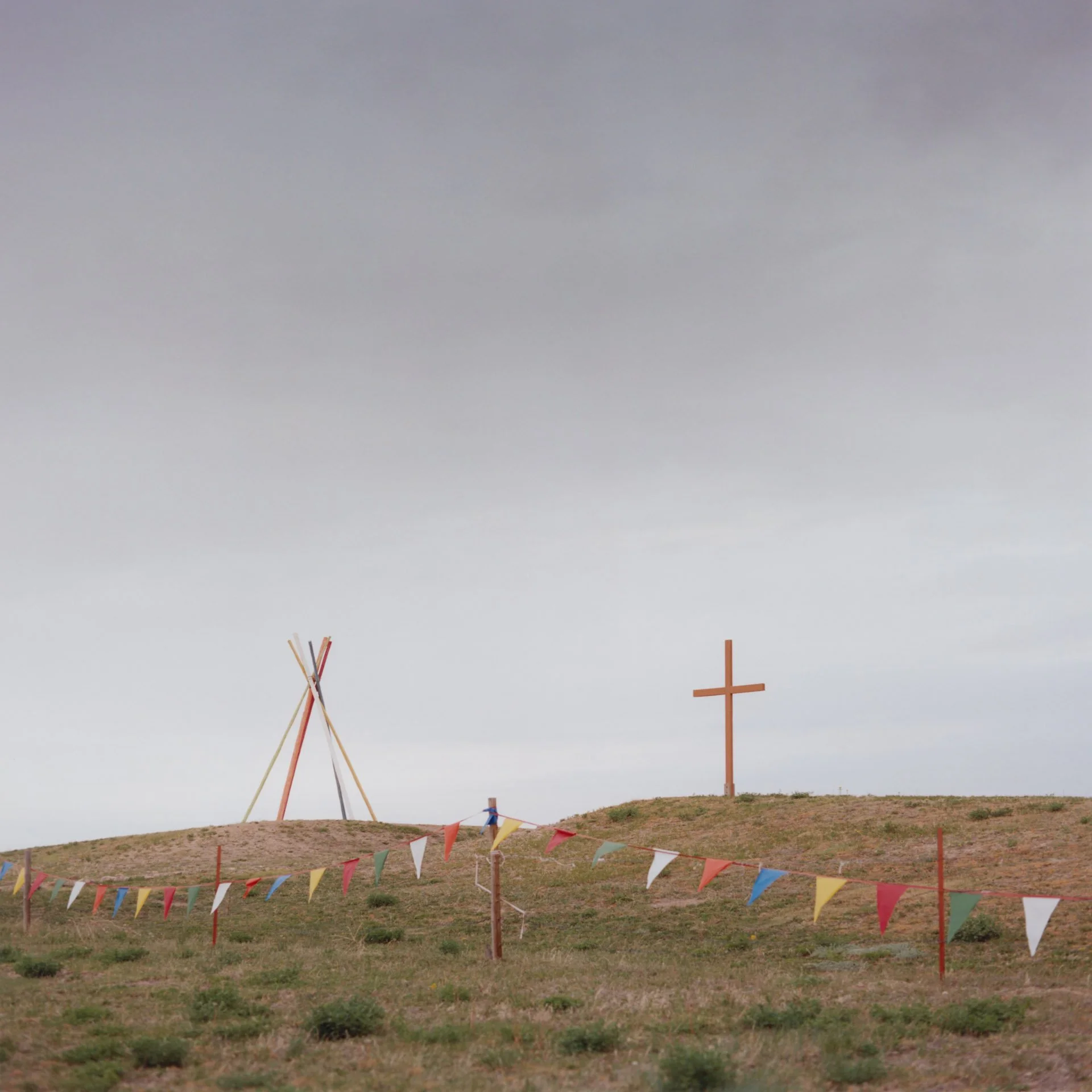 Wooden structures in the shape of a tipi and a cross sit by the side of a road in Pine Ridge, South Dakota. A visual depiction of the crossroads between two worlds of Christianity and traditionalism.