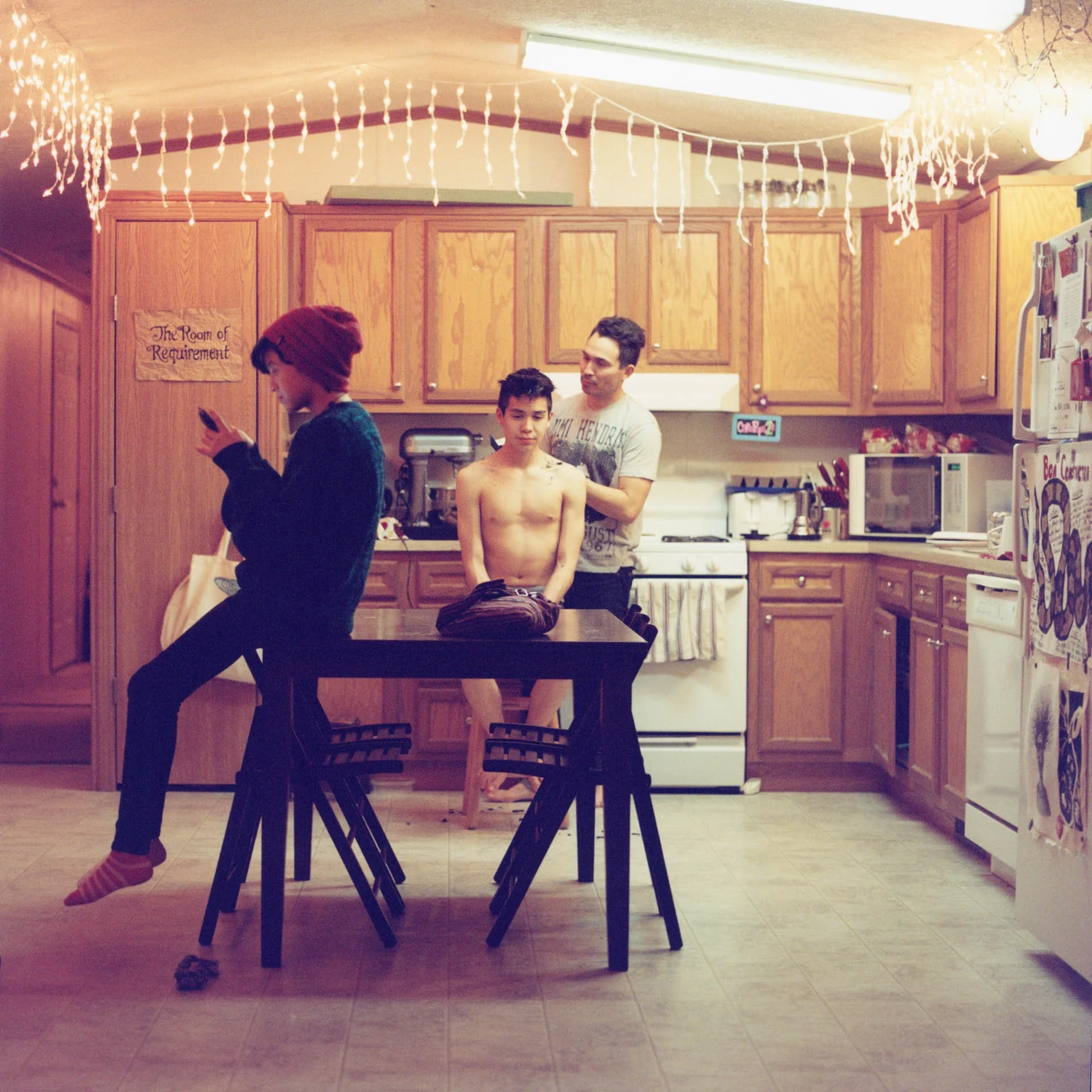 Scott Jr. receives a haircut from his dad while his kid sister hangs out with them in the kitchen.  They are a musical family that reside on the Pine Ridge Indian Reservation and travel around Indian Country performing in their family band.