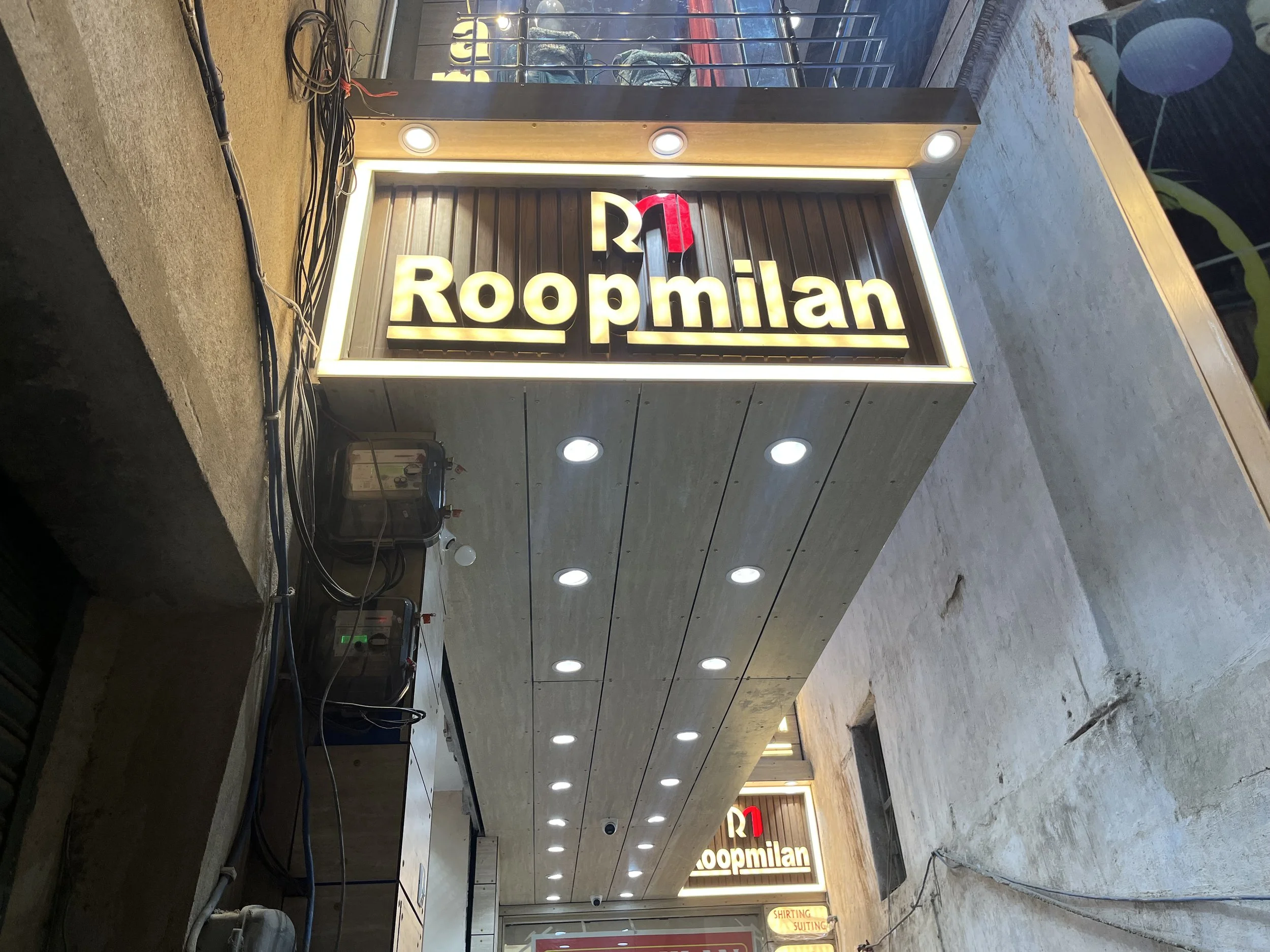 Close-up of a sign for Roopmilan MK, a clothing store for shirt and suit fitting and tailoring, with bright backlit lettering and a red number 1 logo, mounted on a modern exterior wall with recessed lighting.