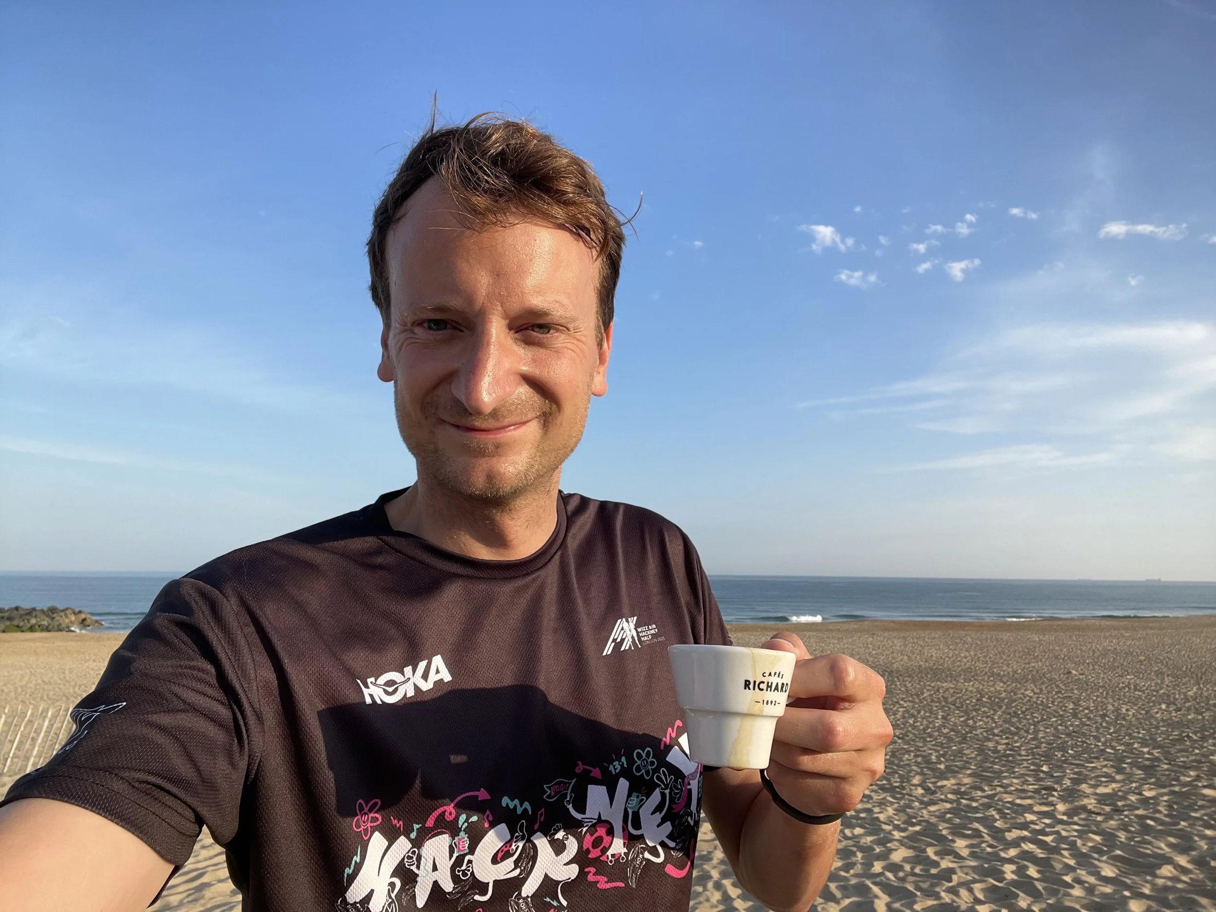 A man smiling at the beach holding a coffee cup, with a sandy shoreline, ocean waves, and blue sky in the background.