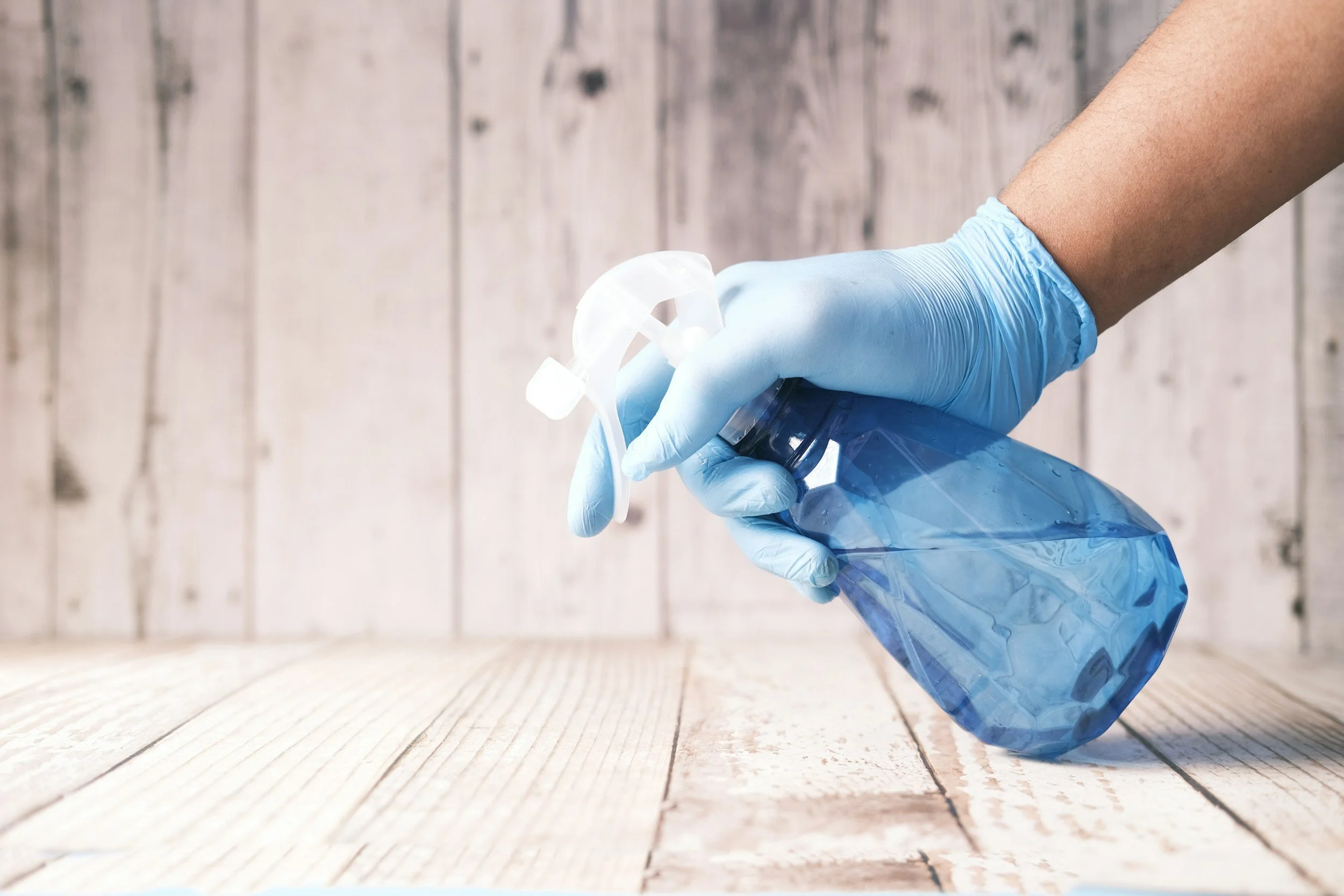 A person wearing a blue glove holding a plastic spray bottle on a wooden surface with a wooden panel background.
