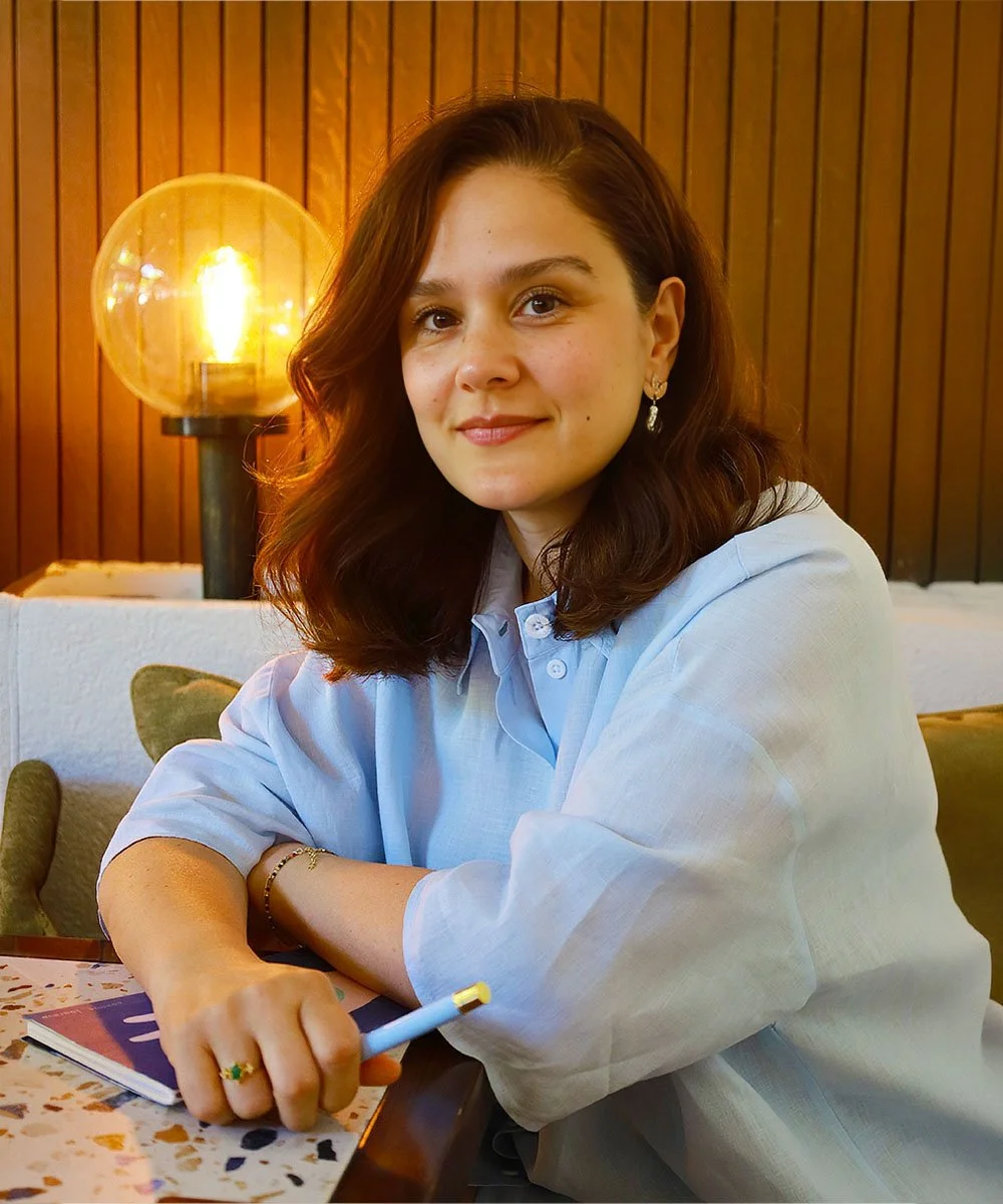 Krema rengi bir koltuk ve arka planda ahşap panel duvarın önünde oturan genç kadın, mavi gömlek giymiş, kahverengi saçlı, gülümseyen, elinde kalem tutuyor, masasında defter ve kitap var, arka planda yuvarlak turuncu ışık lambası açık.