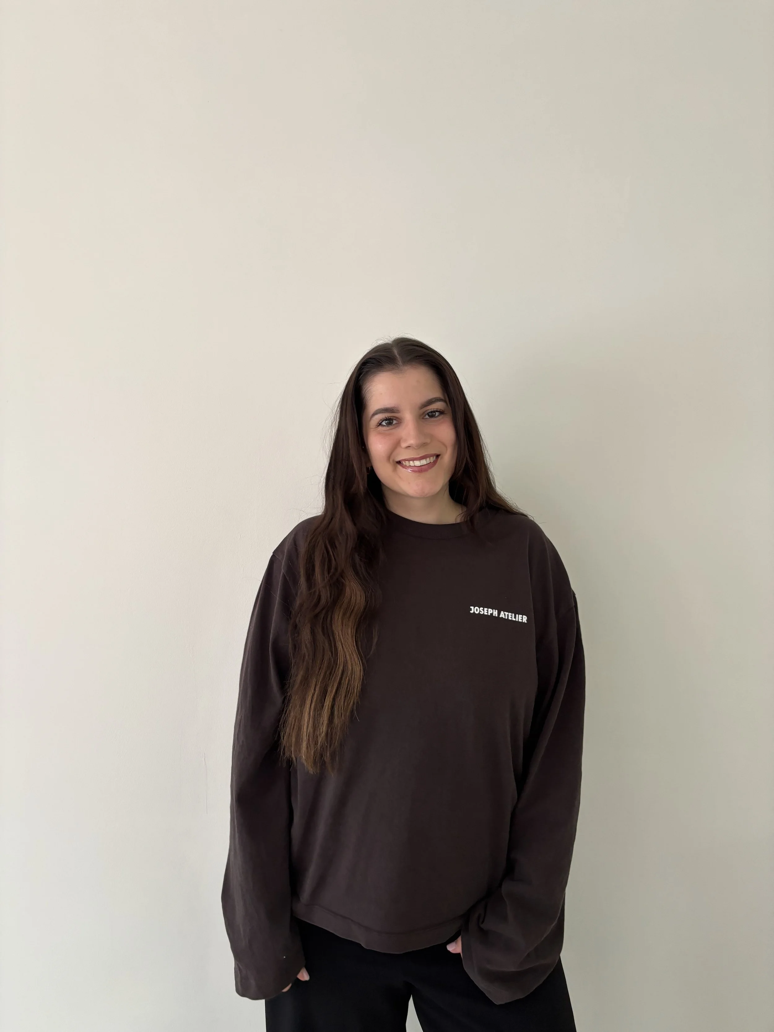 Junge Frau, Nell Meißner, mit braunen, lockigen Haaren, die ein dunkelbraunes Sweatshirt trägt, das die Worte 'JOSEPH ATELIER' auf der linken Brustseite hat, steht vor einer hellen Wand und lächelt.
