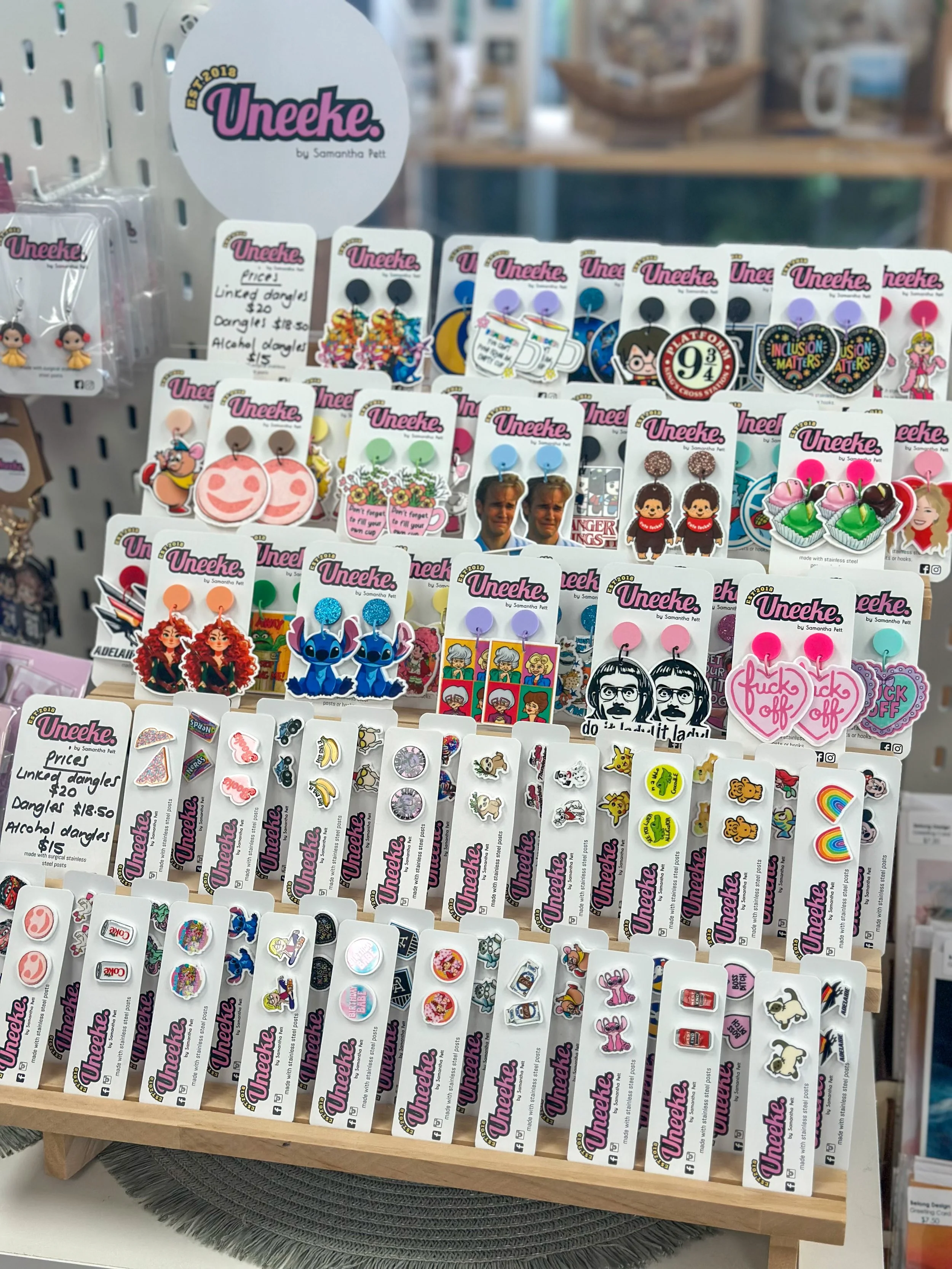 Display of colorful earrings and pins with various designs on a wooden stand at a store. The earrings include characters, icons, and humorous messages, with branded tags reading 'Uneeke'.