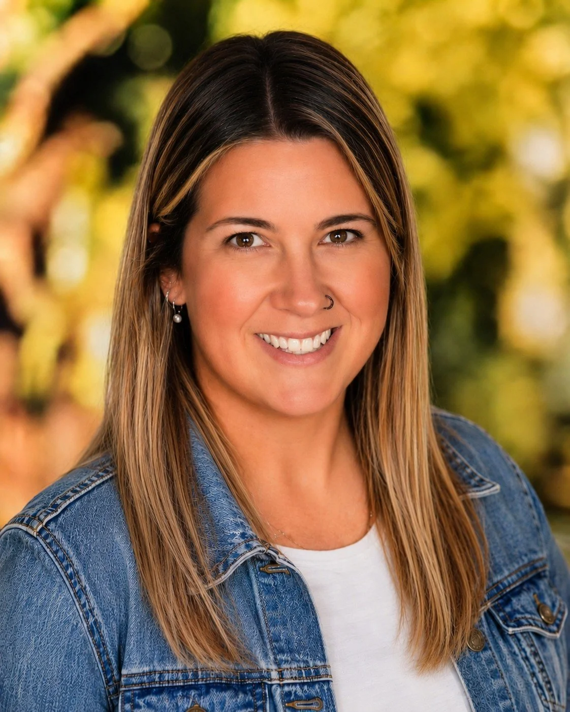 A woman with straight, shoulder-length brown hair with blonde highlights, smiling, wearing a denim jacket and pearl earrings, outdoors with blurred trees and sunlight in the background.
